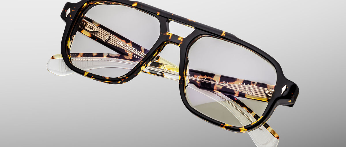 The Jacques Marie Mage Evans eyeglasses feature an oversized tortoiseshell acetate frame, clear lenses, and gold temple accents, presented against a light gray background.