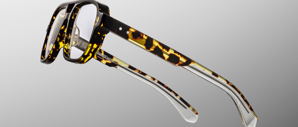 The Jacques Marie Mage Evans optical glasses showcase a handcrafted, oversized rectangular acetate frame in tortoiseshell with clear lower arms, set against a light gray background.