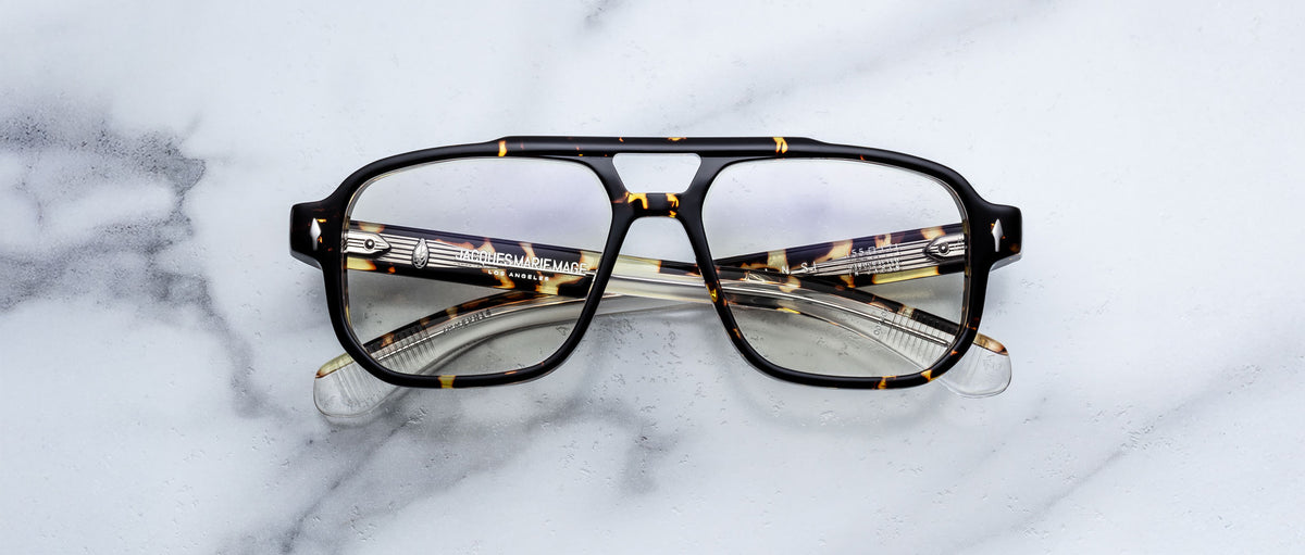 The Jacques Marie Mage Evans eyeglasses, featuring an oversized tortoiseshell acetate frame and clear lenses, rest on a white marble surface.