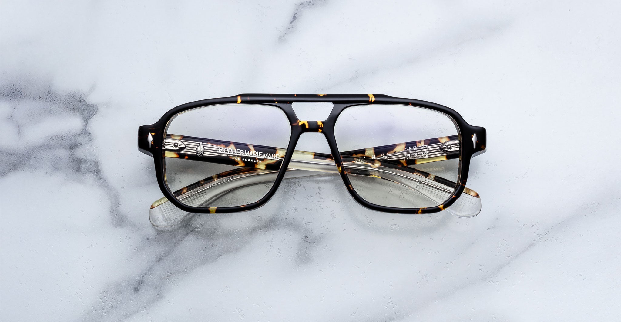 The Jacques Marie Mage Evans eyeglasses, featuring an oversized tortoiseshell acetate frame and clear lenses, rest on a white marble surface.