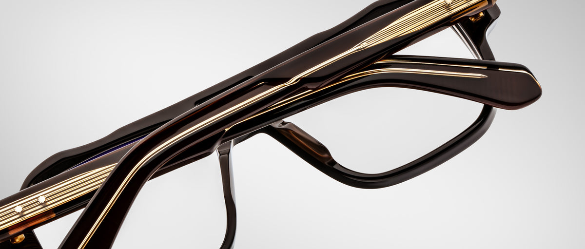 Close-up of folded Jacques Marie Mage Evans eyeglasses featuring black and gold oversized acetate frames, viewed from the side against a plain light background.