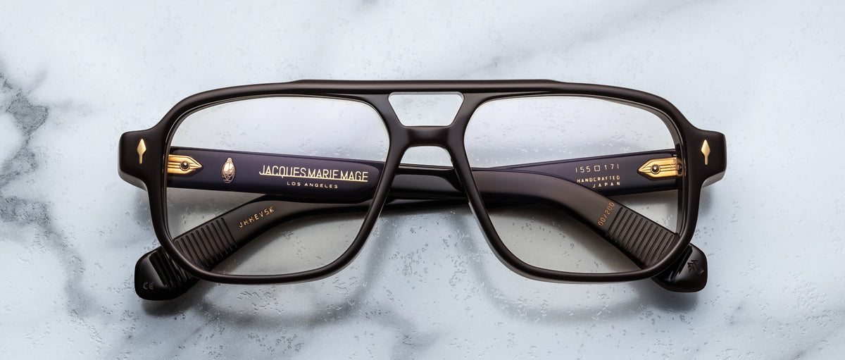 The Jacques Marie Mage Evans are thick-rimmed rectangular eyeglasses with black acetate frames and gold detailing by Jacques Marie Mage, displayed on a light marble surface.