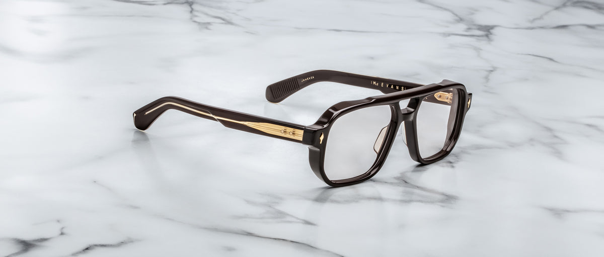 The Jacques Marie Mage Evans eyeglasses, featuring a bold rectangular acetate frame in black and gold, rest elegantly on a white marble surface.