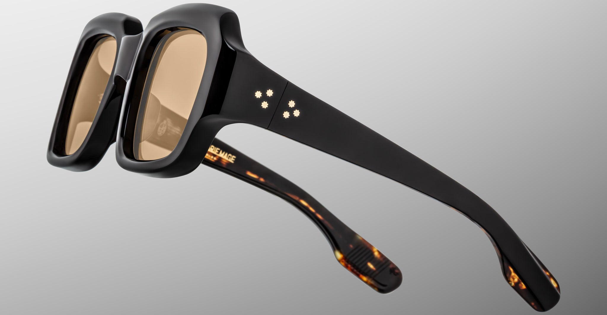 The Jacques Marie Mage Evelyn features chunky black frames with square brown-tinted lenses, silver rivets on the hinges, and subtle tortoiseshell detailing inside the arms, displayed against a gray gradient background.