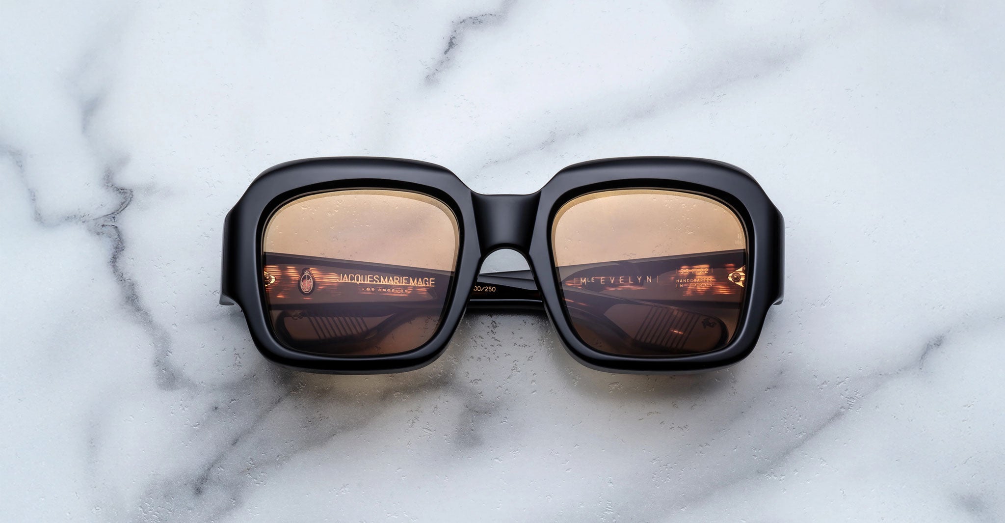 The Jacques Marie Mage Evelyn sunglasses, featuring a square black frame and brown-tinted lenses, rest face up on a white marble surface with small water droplets visible on the bold, thick eyewear.