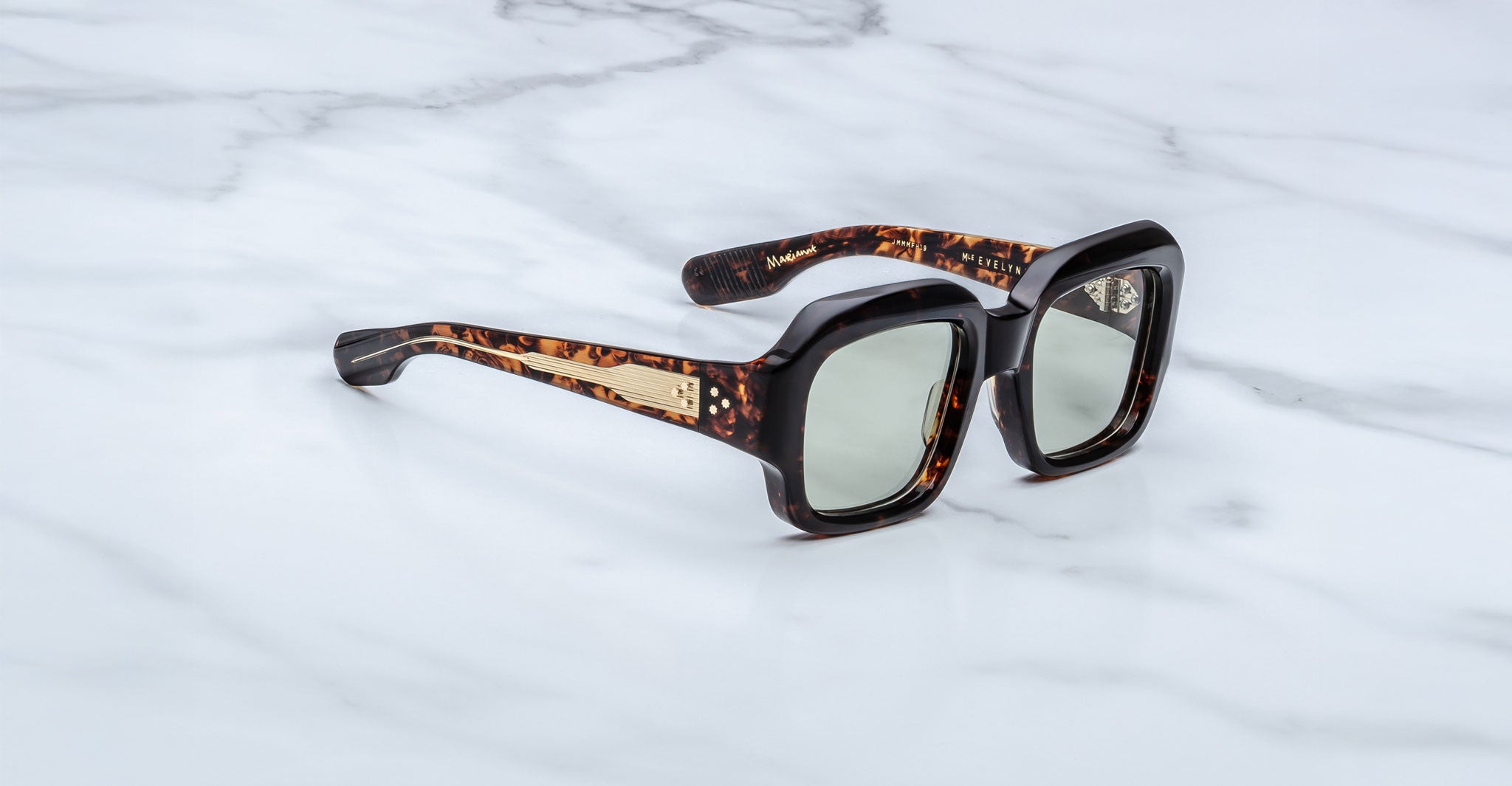 The Jacques Marie Mage Evelyn sunglasses feature a rectangular tortoiseshell design with thick frames and gold-accented arms, displayed on a white marble surface.