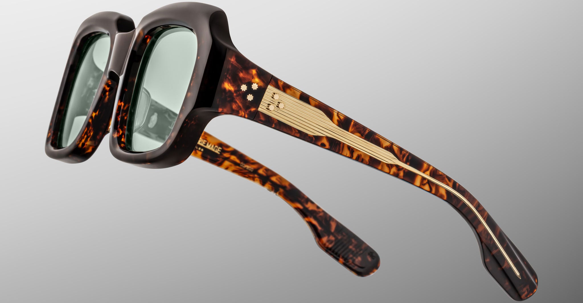 The Jacques Marie Mage Evelyn sunglasses feature thick tortoiseshell rectangular frames, green tinted lenses, and gold metallic accents with a small decorative detail near the hinge.