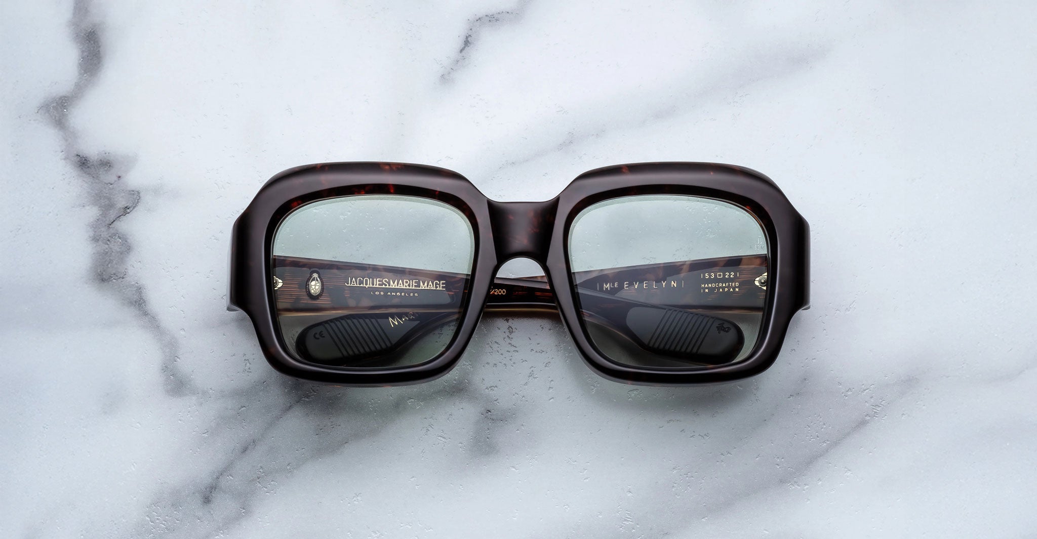A pair of Jacques Marie Mage Evelyn eyeglasses featuring large, square, dark frames with thick rims rests face-up on a white marble surface, with the lens reflections revealing text inside the arms.