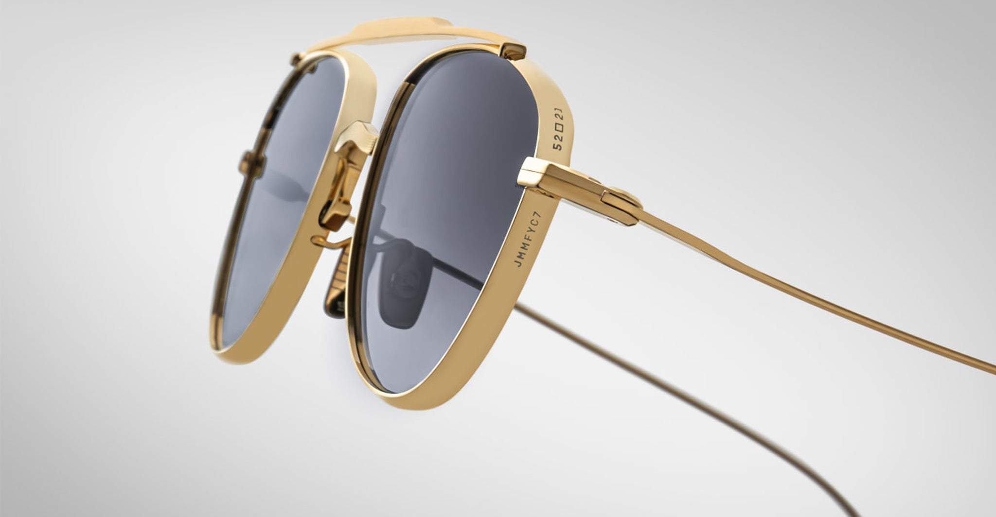 Close-up of Jacques Marie Mage Flynn sunglasses: gold frames, dark tinted lenses, unique double-bridge design, and thin metal arms set against a plain light gray background.