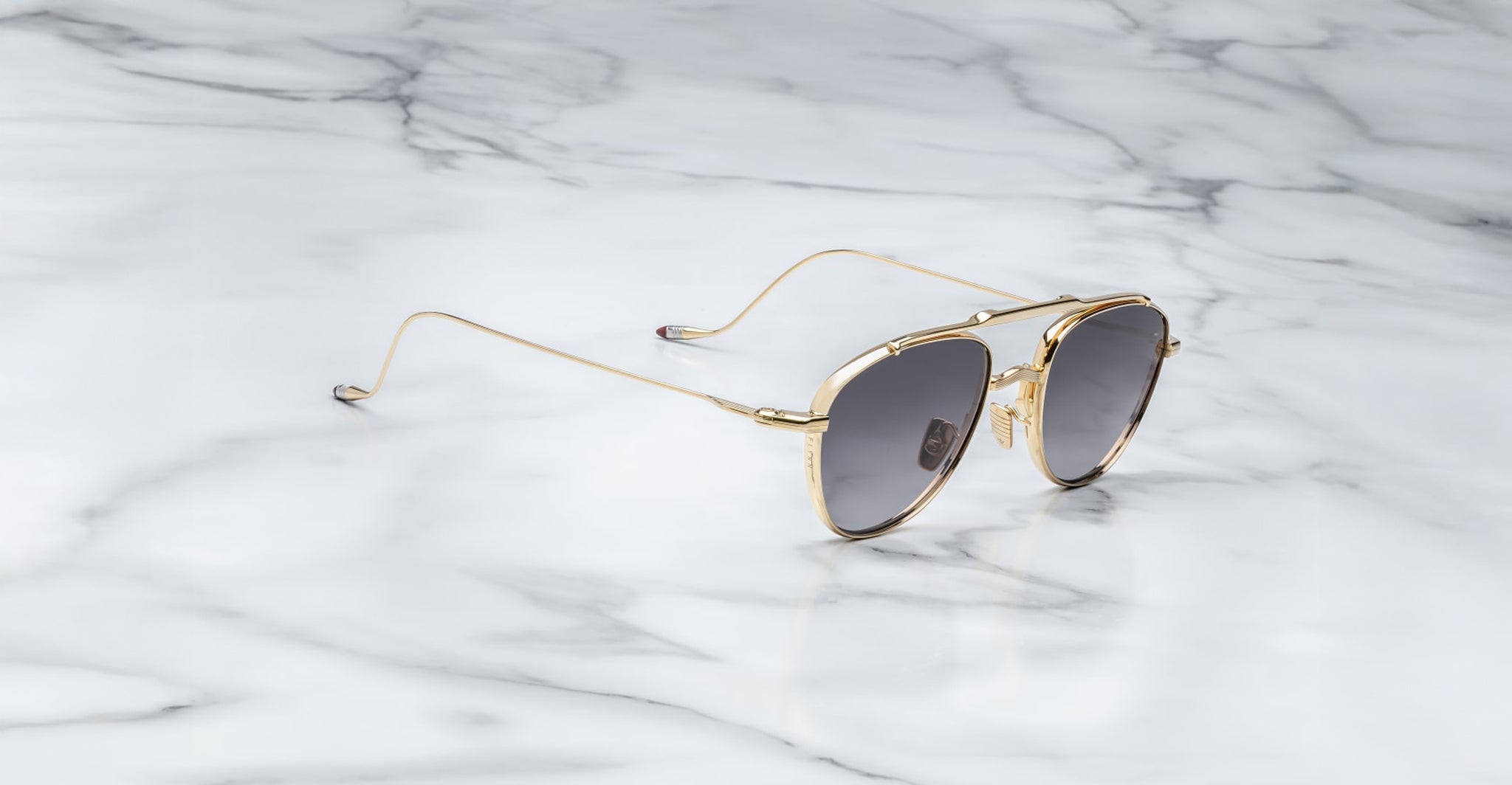 The Jacques Marie Mage Flynn sunglasses, featuring gold frames and dark reflective lenses, are displayed at an angle on a polished white marble surface with gray veining, showcasing their sleek, thin metal arms.