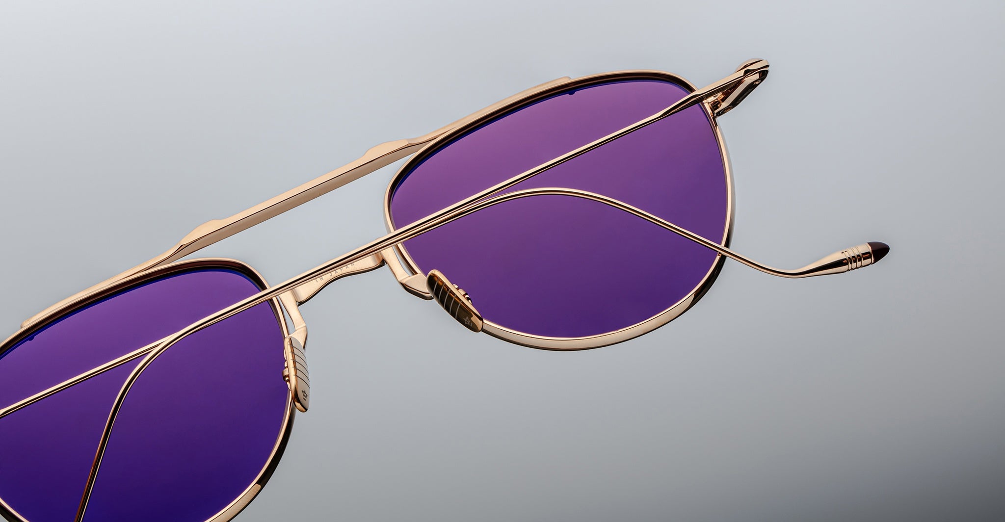 A close-up of Jacques Marie Mage Flynn gold-framed aviator sunglasses with purple-tinted lenses, positioned at an angle on a light gray background.