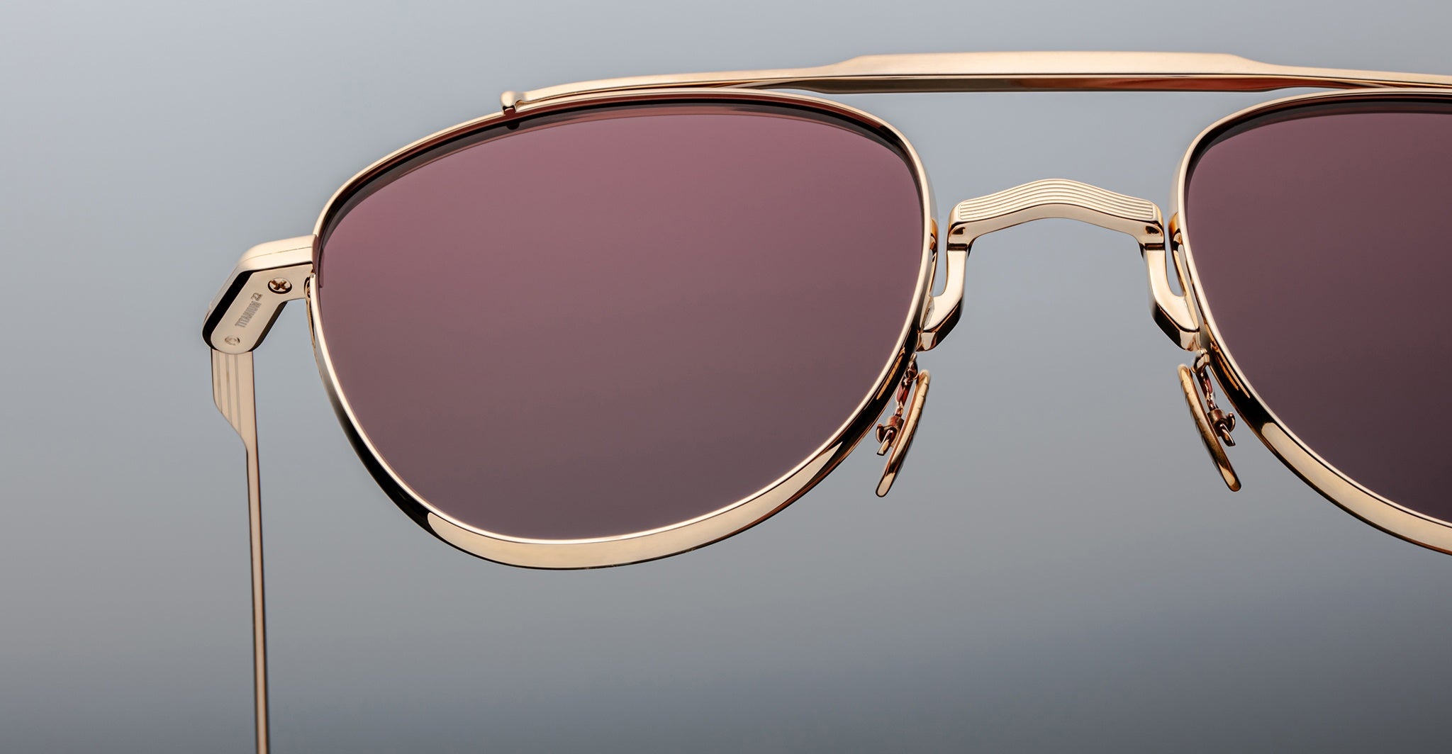 Close-up of Jacques Marie Mage Flynn sunglasses with gold frames, pink-tinted lenses, and a double bridge design, set against a plain gray background.