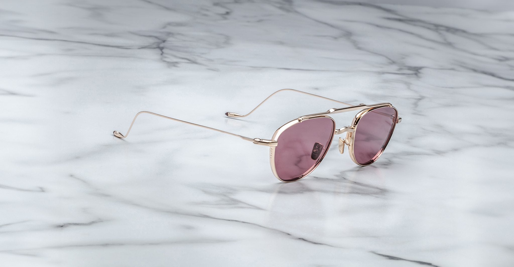 A pair of Jacques Marie Mage Flynn gold-framed sunglasses with pink-tinted lenses rests on a white marble surface.