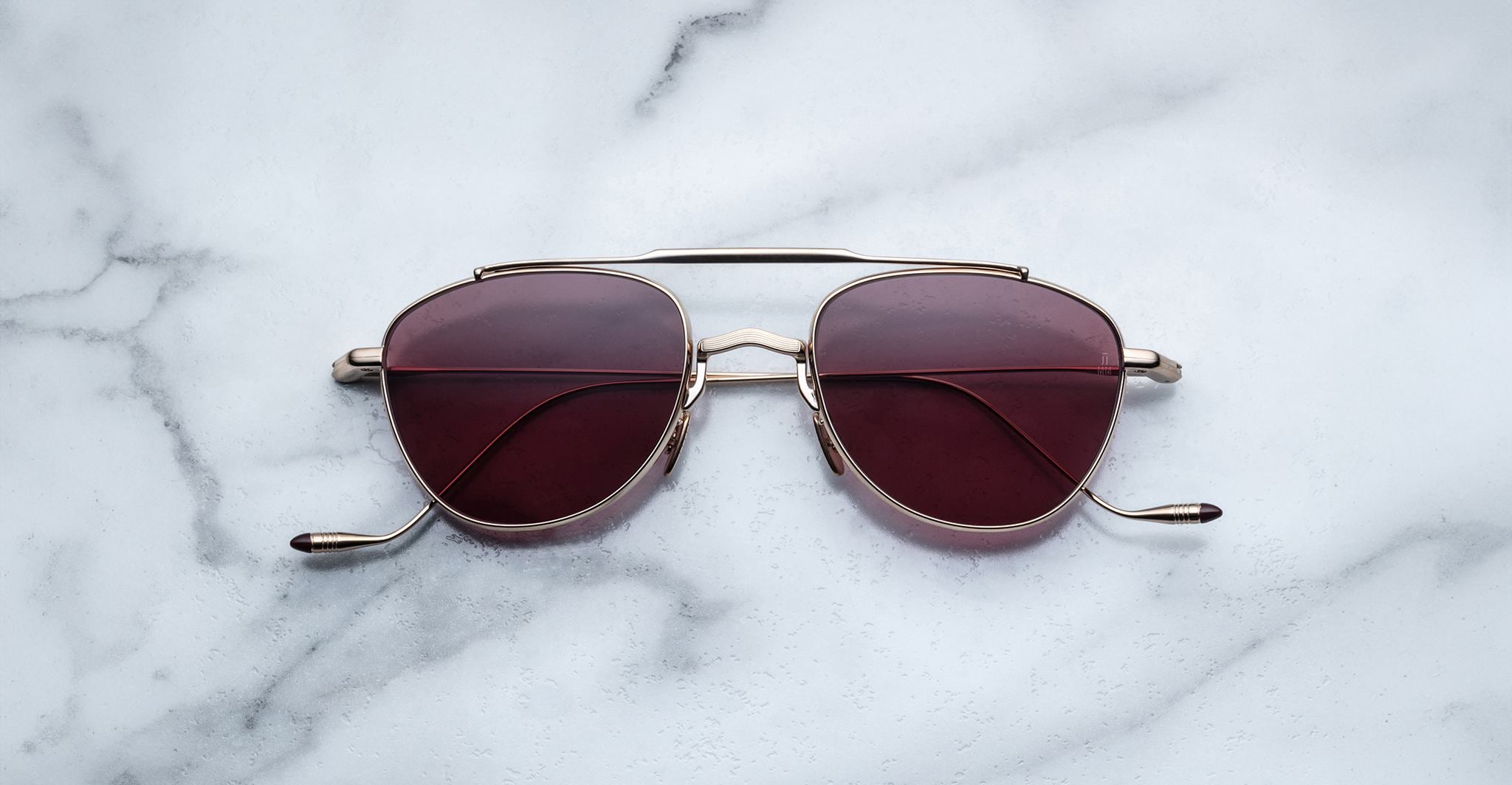 A pair of Jacques Marie Mage Flynn sunglasses with gold frames and dark red lenses rests on a white marble surface.