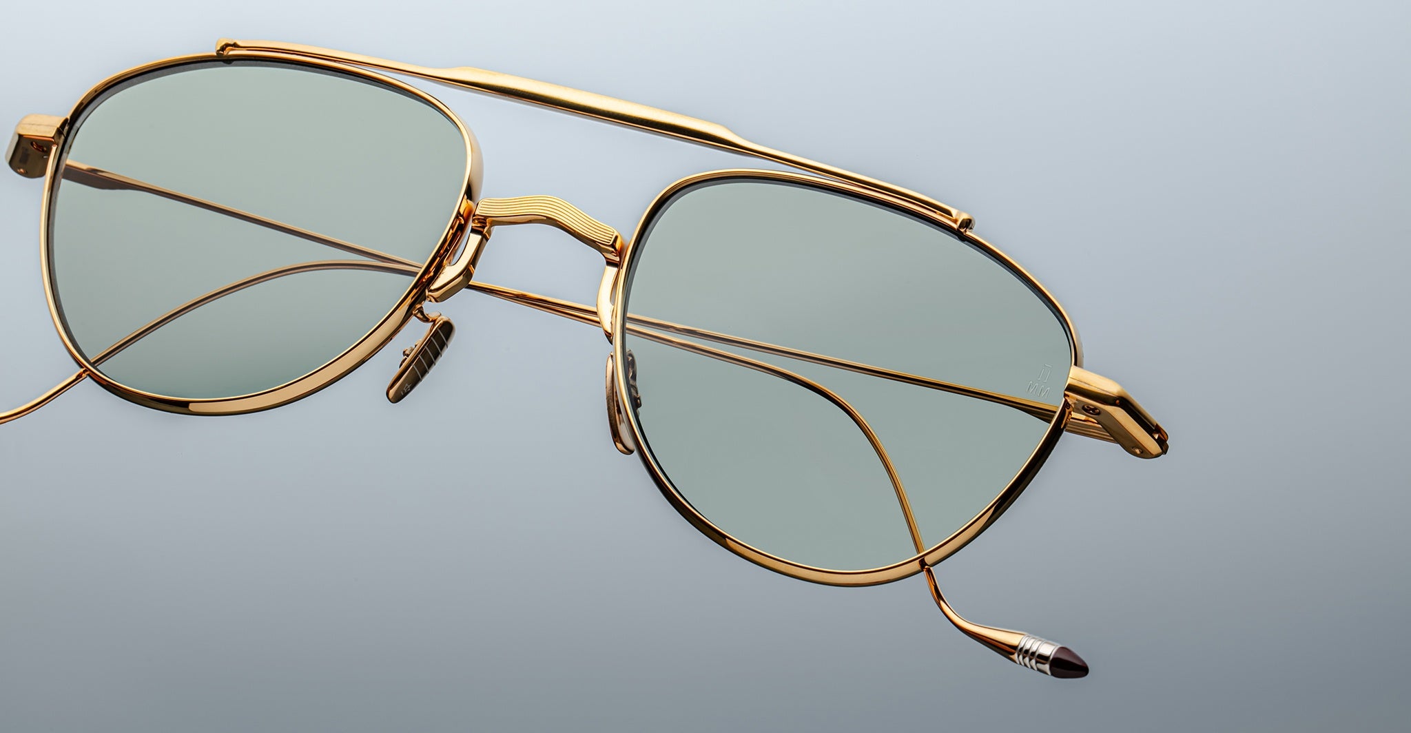 The Jacques Marie Mage Flynn eyeglasses feature gold aviator-style frames and light green tinted lenses, displayed against a plain light gray background.