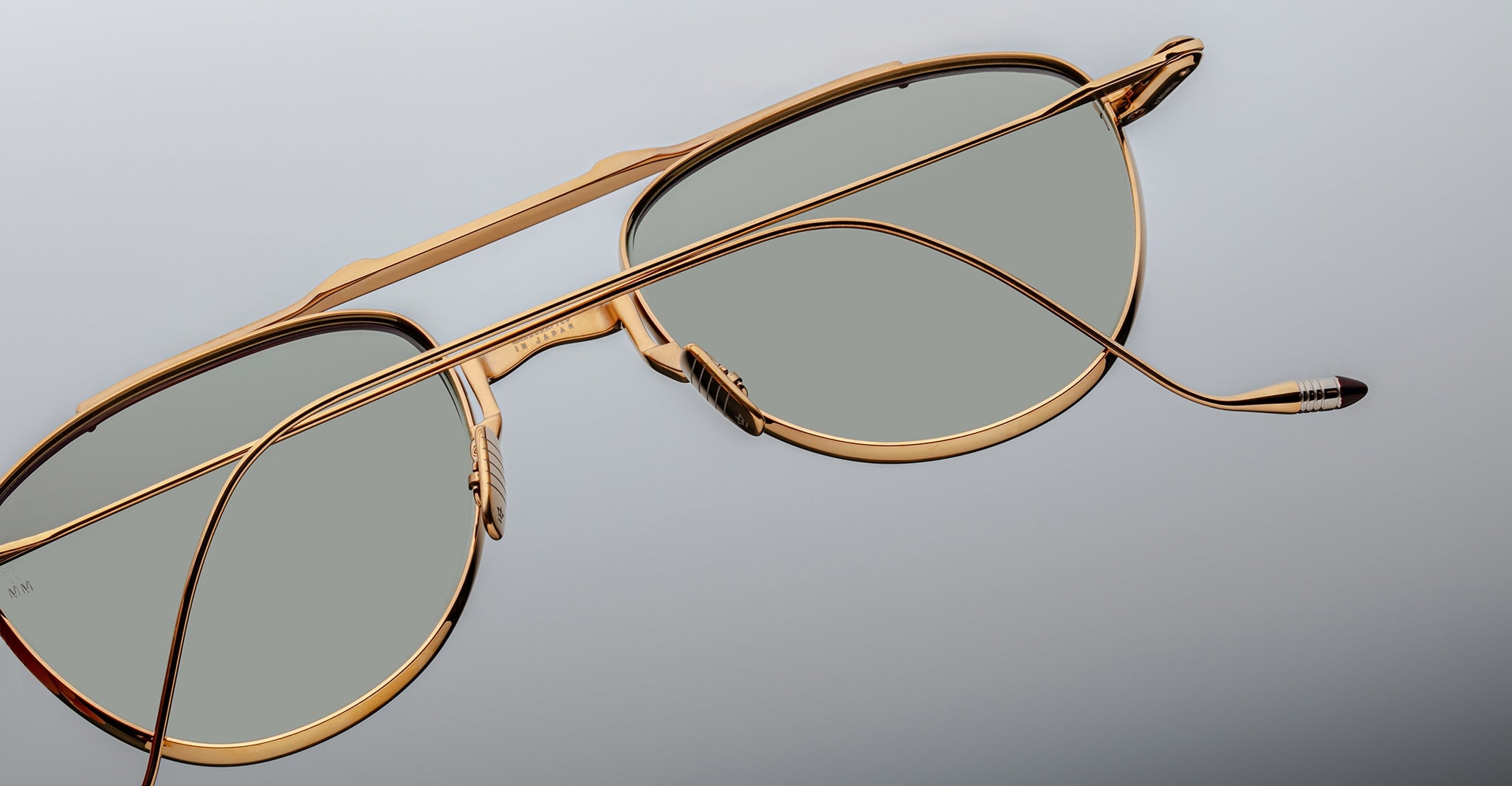 Close-up of Jacques Marie Mage Flynn aviator sunglasses with gold frames and green lenses, placed on a grey surface—one arm extended, the other resting across the lenses.