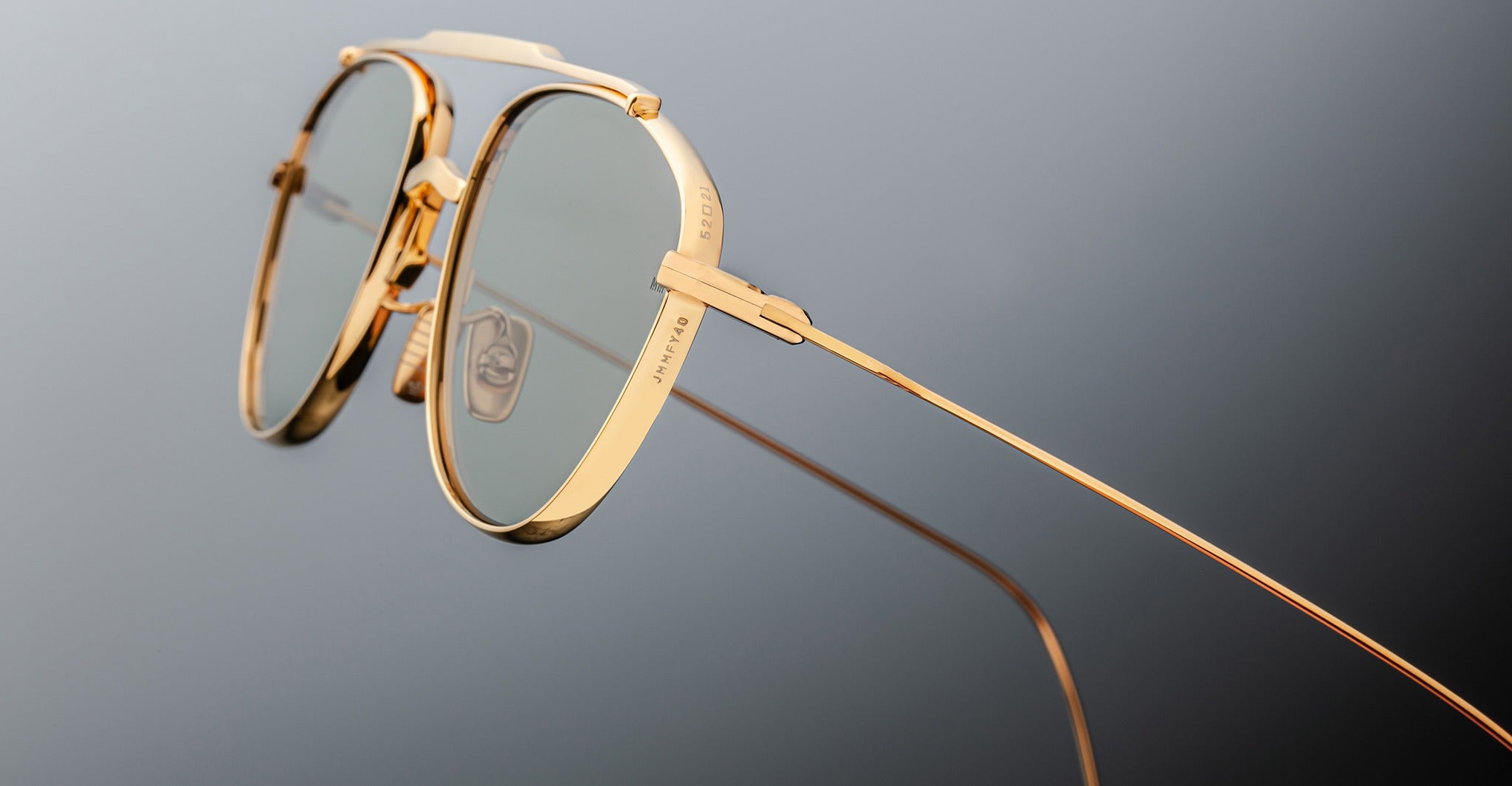Close-up of Jacques Marie Mage Flynn gold-framed aviator eyeglasses with clear lenses, highlighting detailed design and thin temples against a neutral gray background.