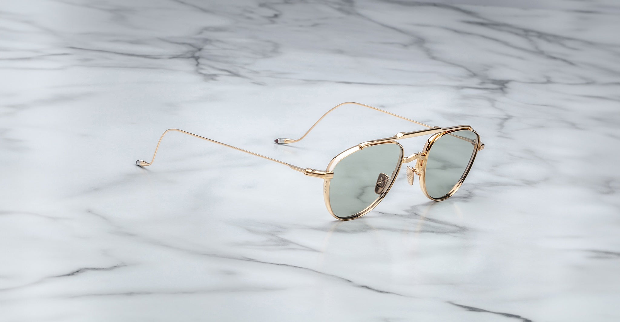 A pair of Jacques Marie Mage Flynn gold-framed aviator sunglasses with green-tinted lenses rests on a white marble surface.