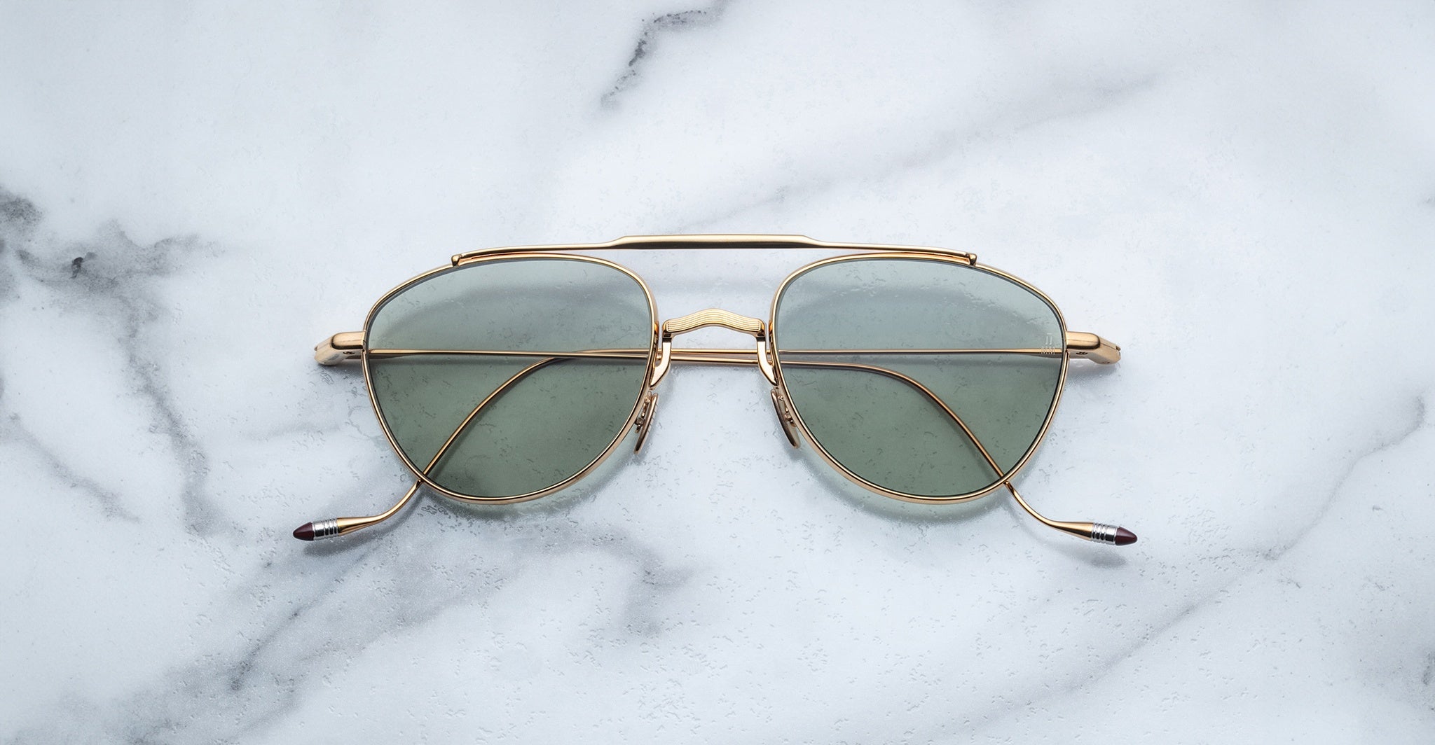 A pair of Jacques Marie Mage Flynn gold-rimmed aviator sunglasses with green-tinted lenses rests on a white marble surface featuring subtle gray veins.