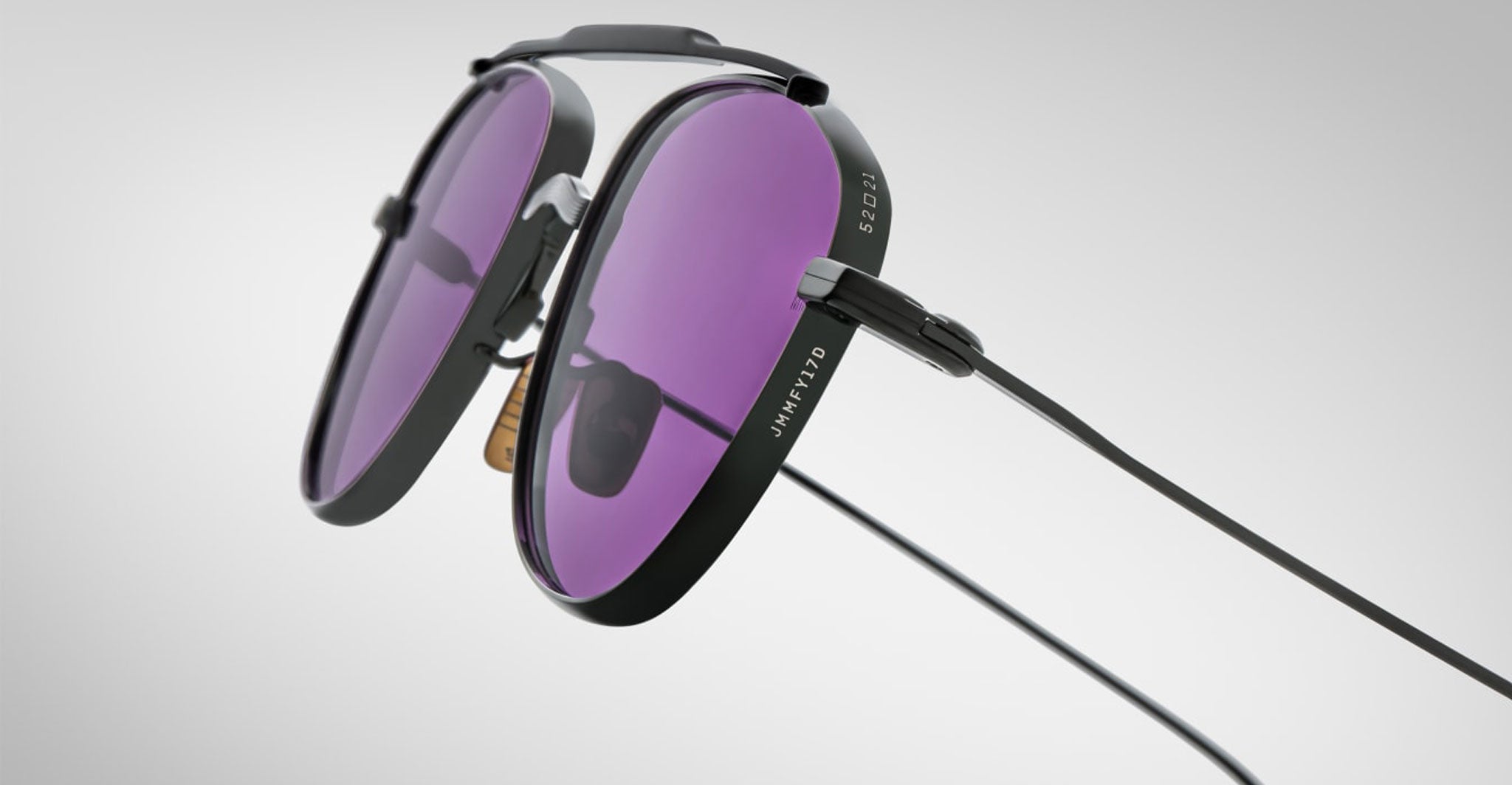 A close-up of Jacques Marie Mage Flynn sunglasses featuring round, purple-tinted lenses, black frames with detachable side shields, and a thin black cord on the temples against a plain light gray background.