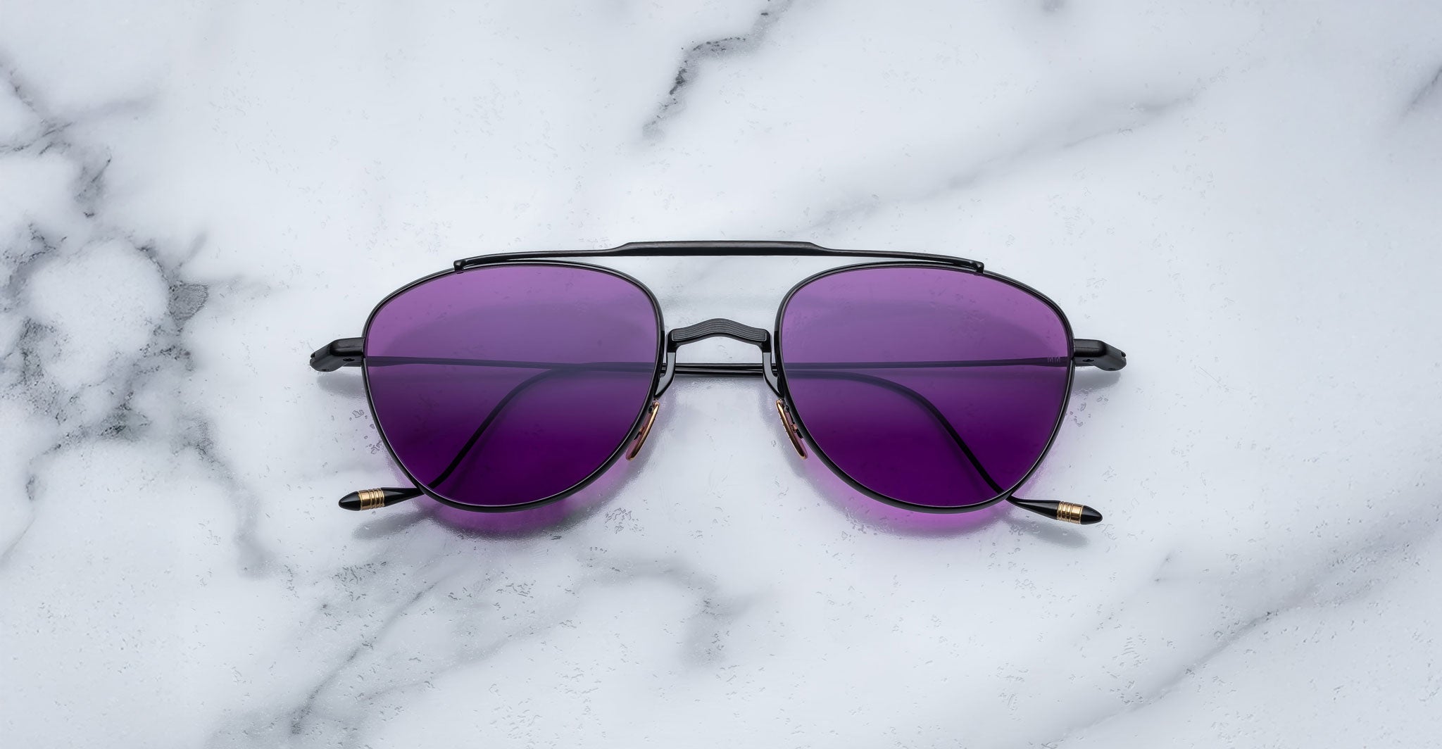A pair of Jacques Marie Mage Flynn sunglasses with black metal frames and purple-tinted lenses rests on a white marble surface.