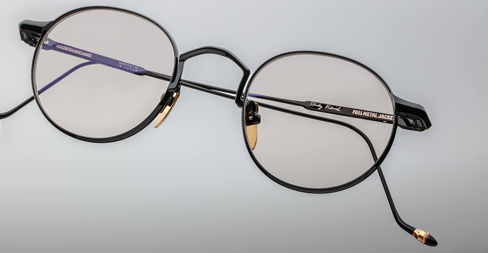 A pair of Jacques Marie Mage Full Metal Jacket round eyeglasses with metal frames, clear lenses, gold nose pad accents, black temple arms, and FULL METAL JACKET inscribed on the right arm.