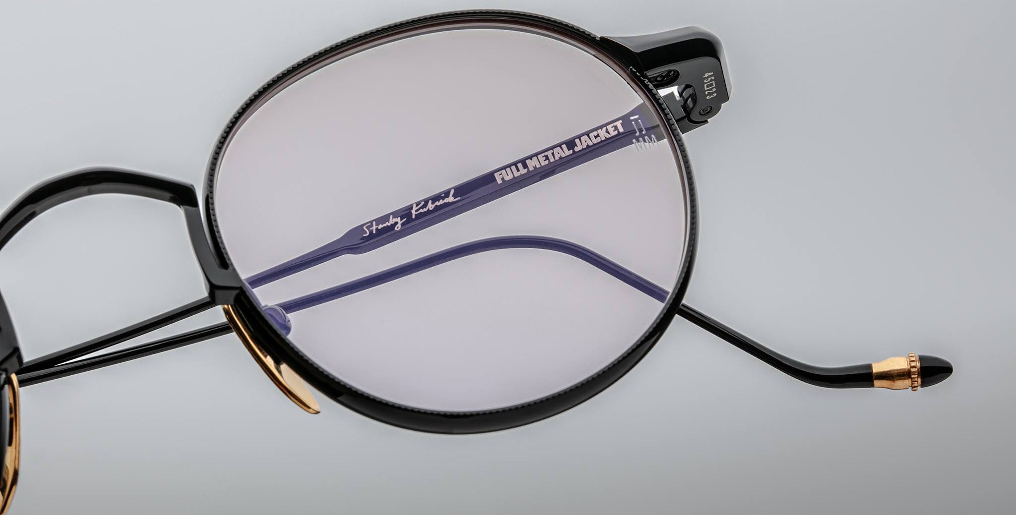 A close-up of Jacques Marie Mage Full Metal Jacket eyeglasses featuring black and gold frames. Through the lens, the blue inner arm displays FULL METAL JACKET and a signature, set against a soft light gray background.