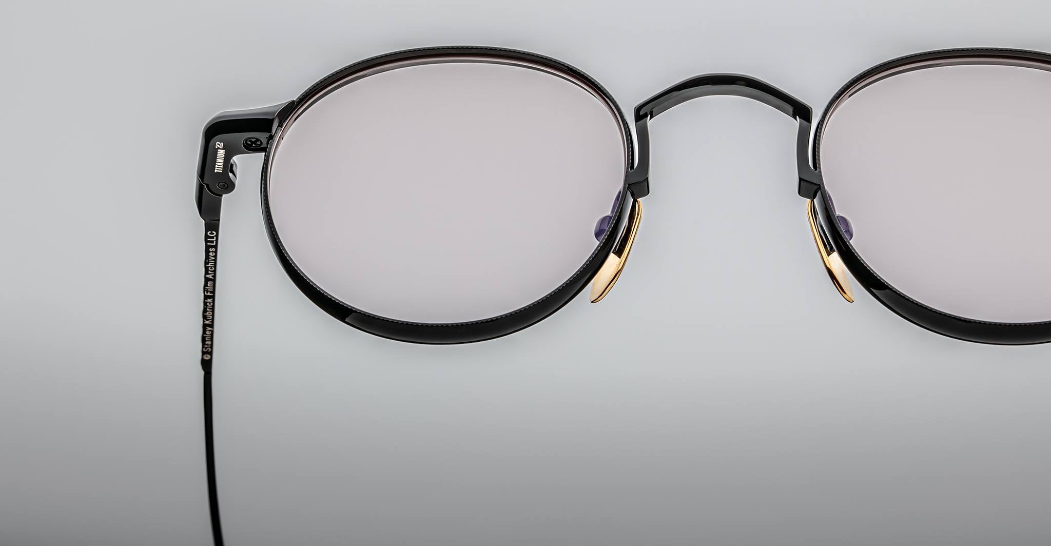 Close-up of Jacques Marie Mage Full Metal Jacket eyeglasses—black round frames with gold accents—set against a plain light gray background.