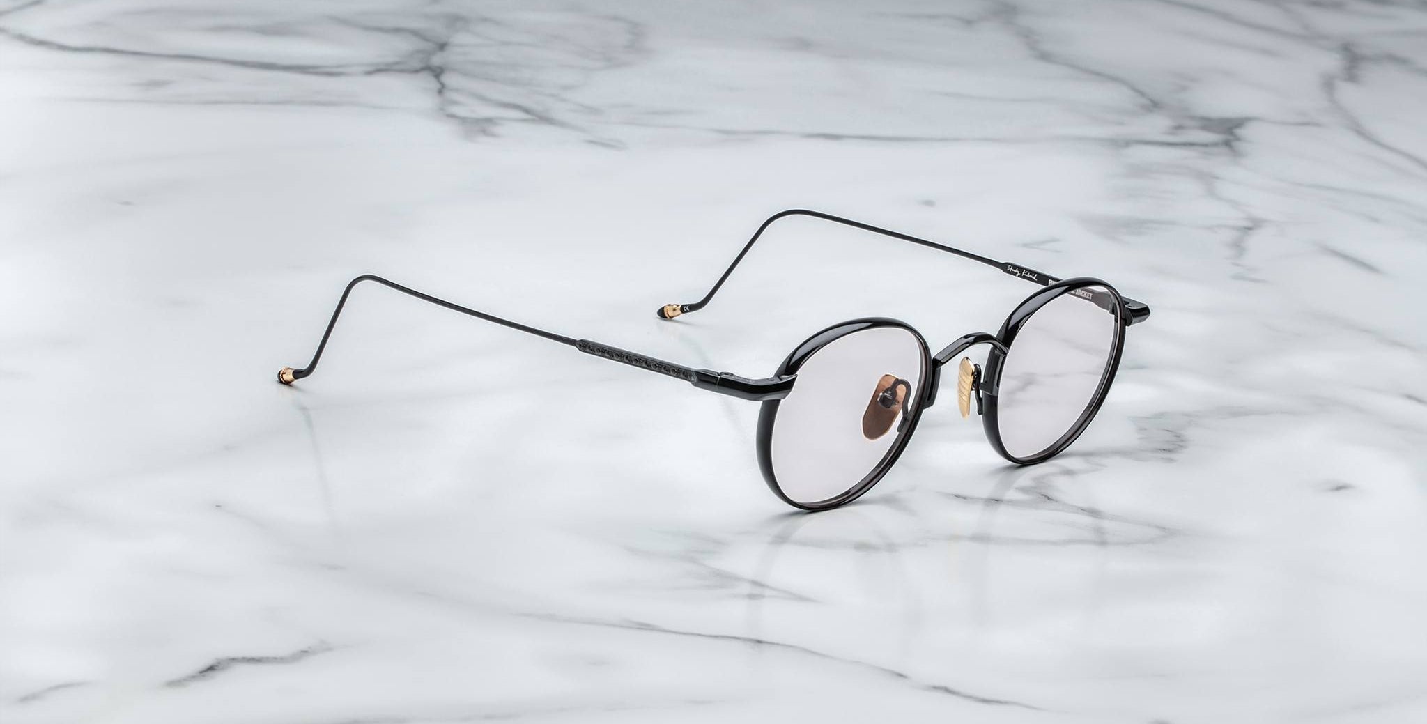 The Jacques Marie Mage Full Metal Jacket eyeglasses, featuring round black rims, thin arms, and gold accents, rest on a white marble surface with gray veining.