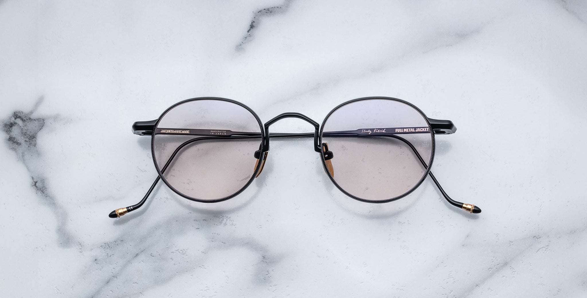 The Jacques Marie Mage Full Metal Jacket glasses, featuring round dark frames and lightly tinted lenses, are displayed on a white marble surface with delicate gray veining.