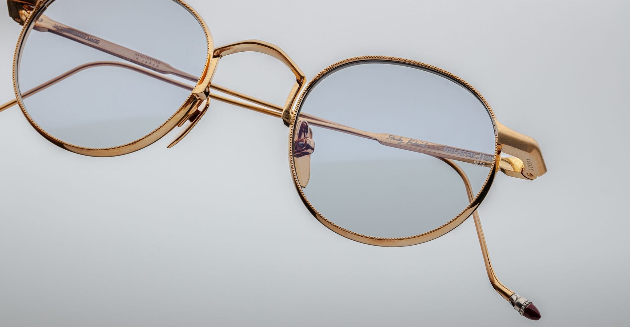 Close-up of Jacques Marie Mage Full Metal Jacket eyeglasses: gold metal frame, light blue round lenses from an angle