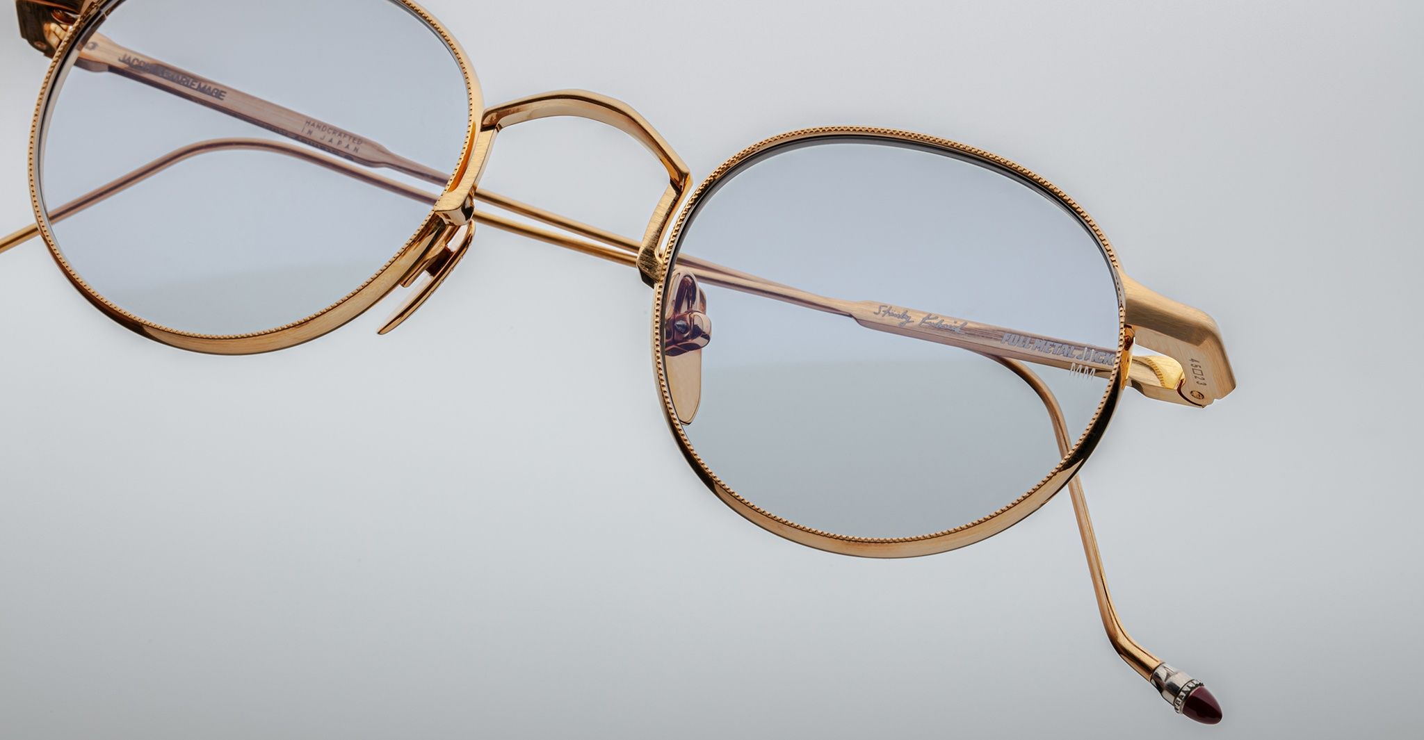 Close-up of Jacques Marie Mage Full Metal Jacket eyeglasses: gold metal frame, light blue round lenses from an angle