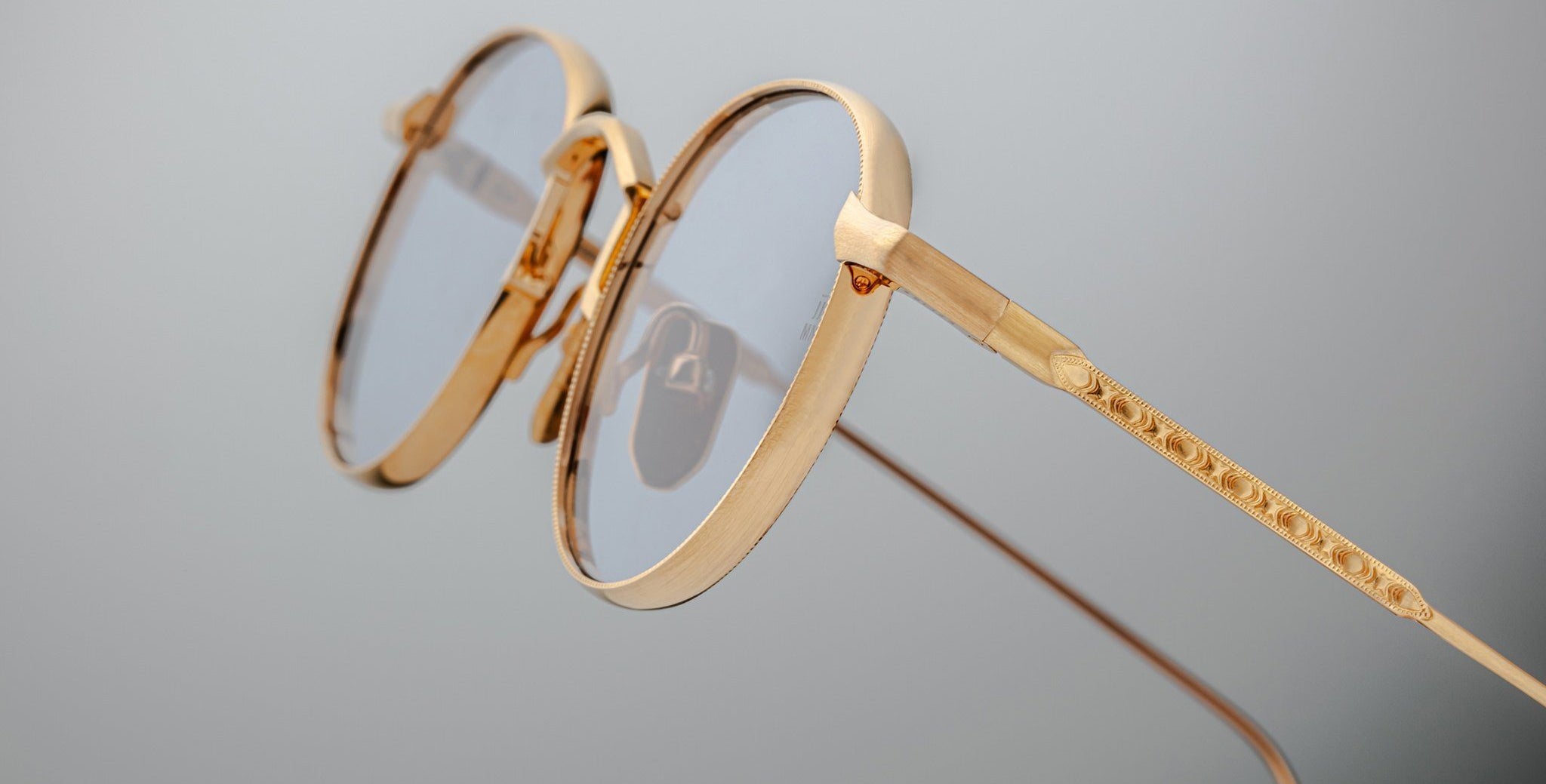 Close-up of Jacques Marie Mage Full Metal Jacket eyeglasses: gold-framed, round with clear lenses, featuring intricate detailing on the arms, set against a plain light gray background.