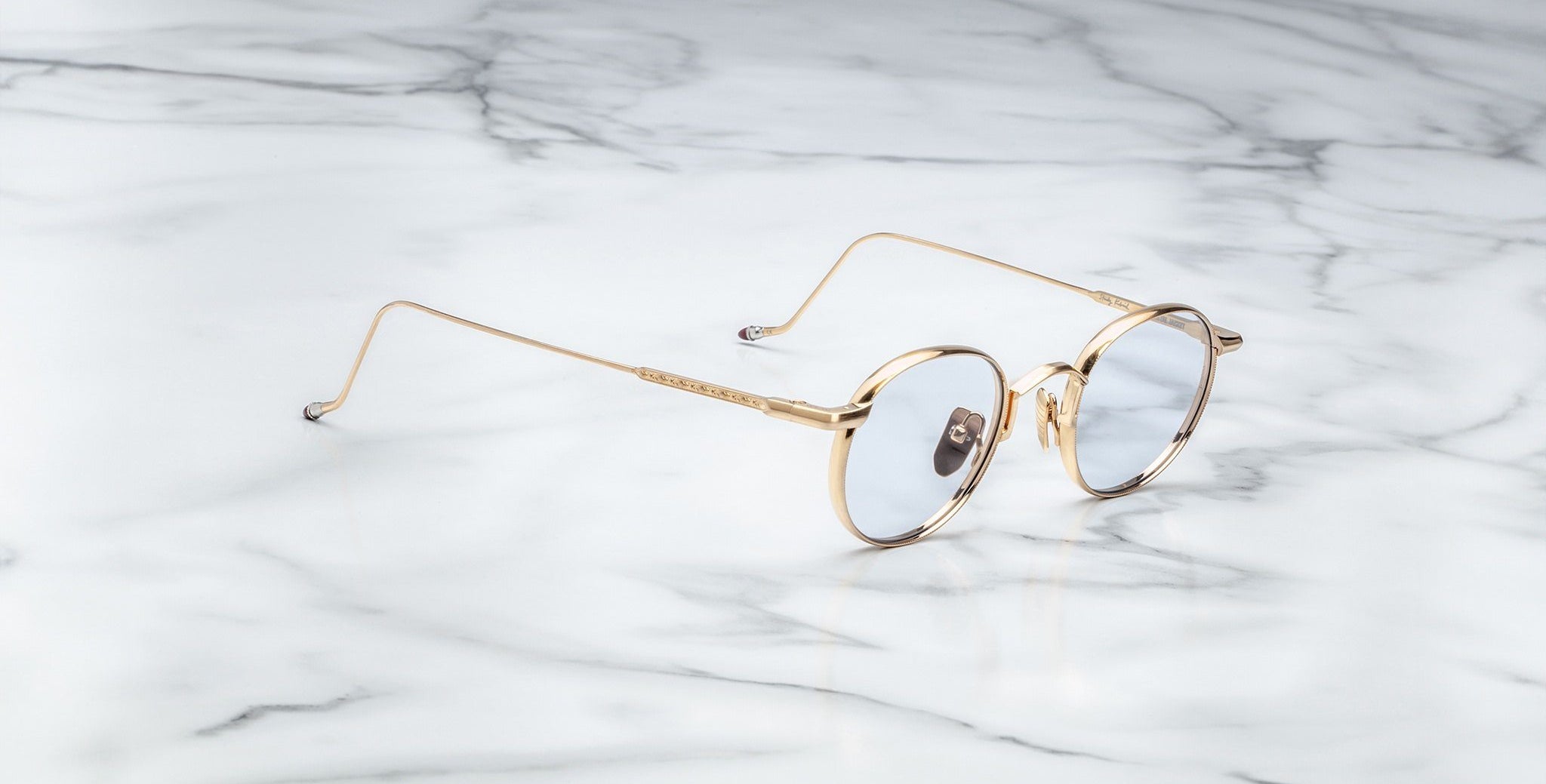 The Jacques Marie Mage Full Metal Jacket eyeglasses, featuring round gold frames with thin arms and clear lenses, rest on a white marble surface with grey veining.