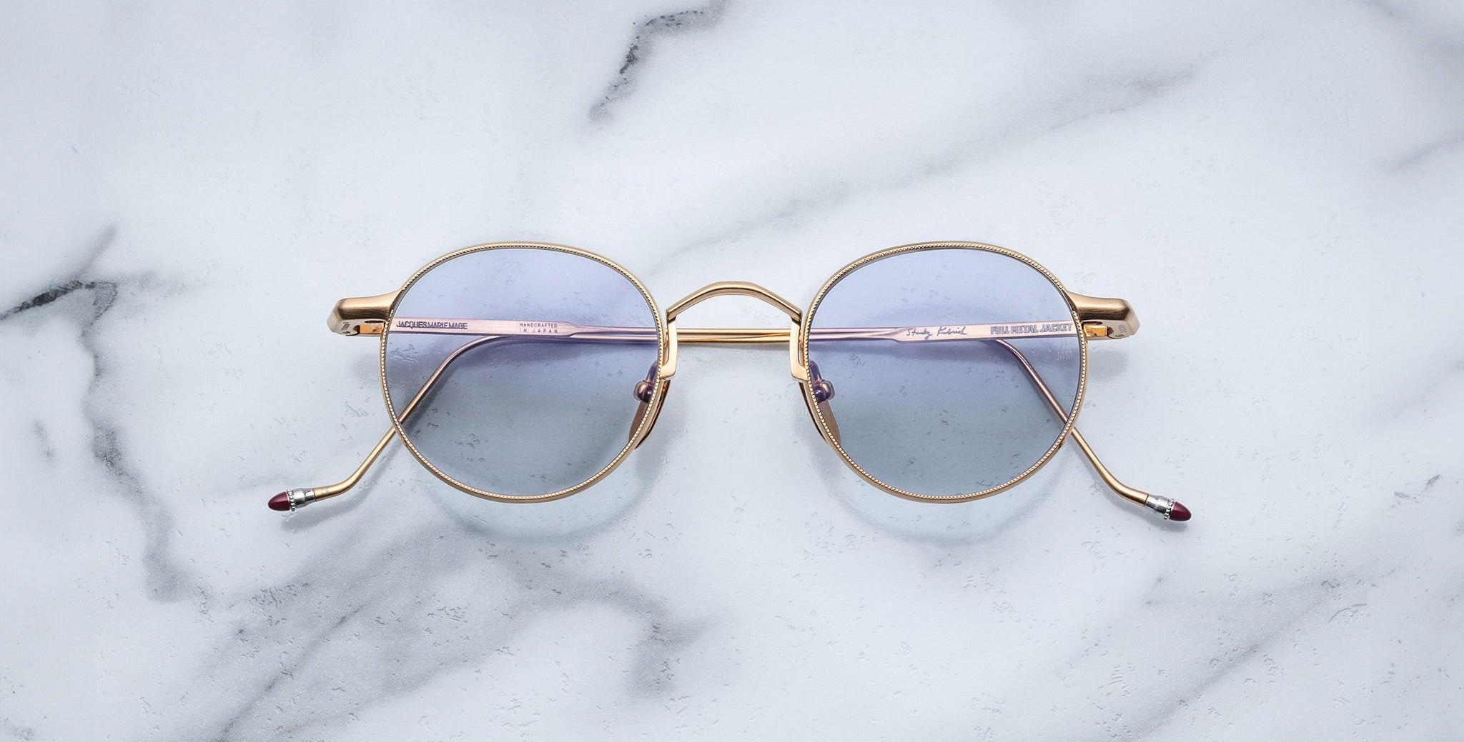 The Jacques Marie Mage Full Metal Jacket sunglasses, featuring round gold frames and light purple-tinted lenses, are displayed on a white marble surface with gray veining.