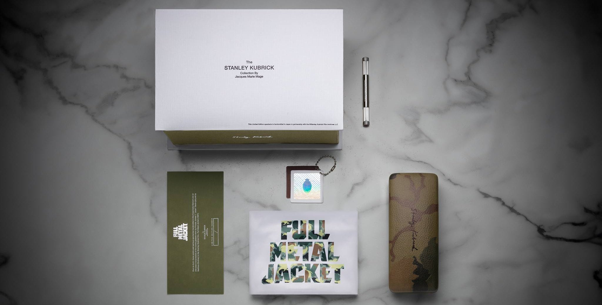 A flat lay of Jacques Marie Mage Full Metal Jacket merchandise on marble features a white box, metal screwdriver, camo glasses case, green envelope, keychain with blue drop, and a graphic card displaying the movie title.