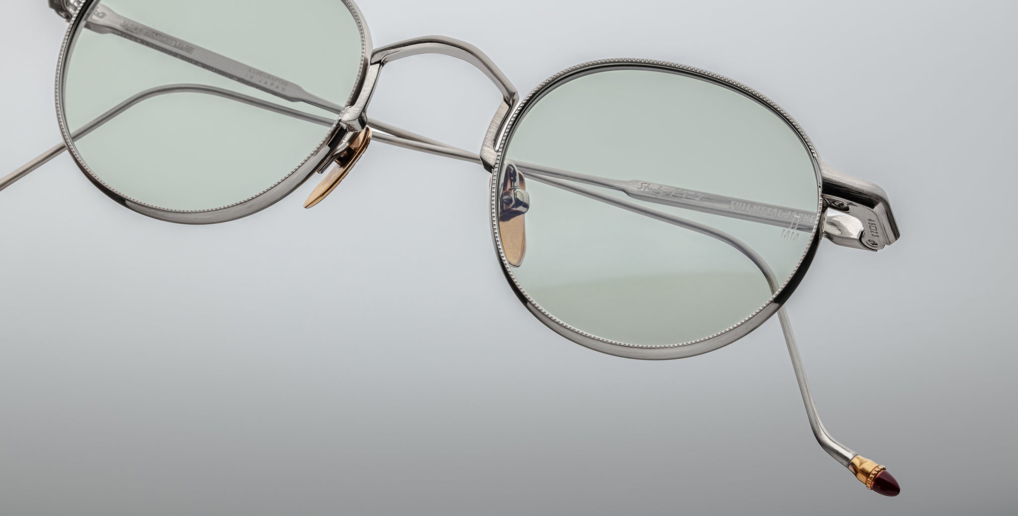 Close-up of Jacques Marie Mage Full Metal Jacket eyeglasses featuring stylish round silver frames, light green lenses, and gold and brown accents on the nose pads and temple tips, set against a smooth light gradient background.
