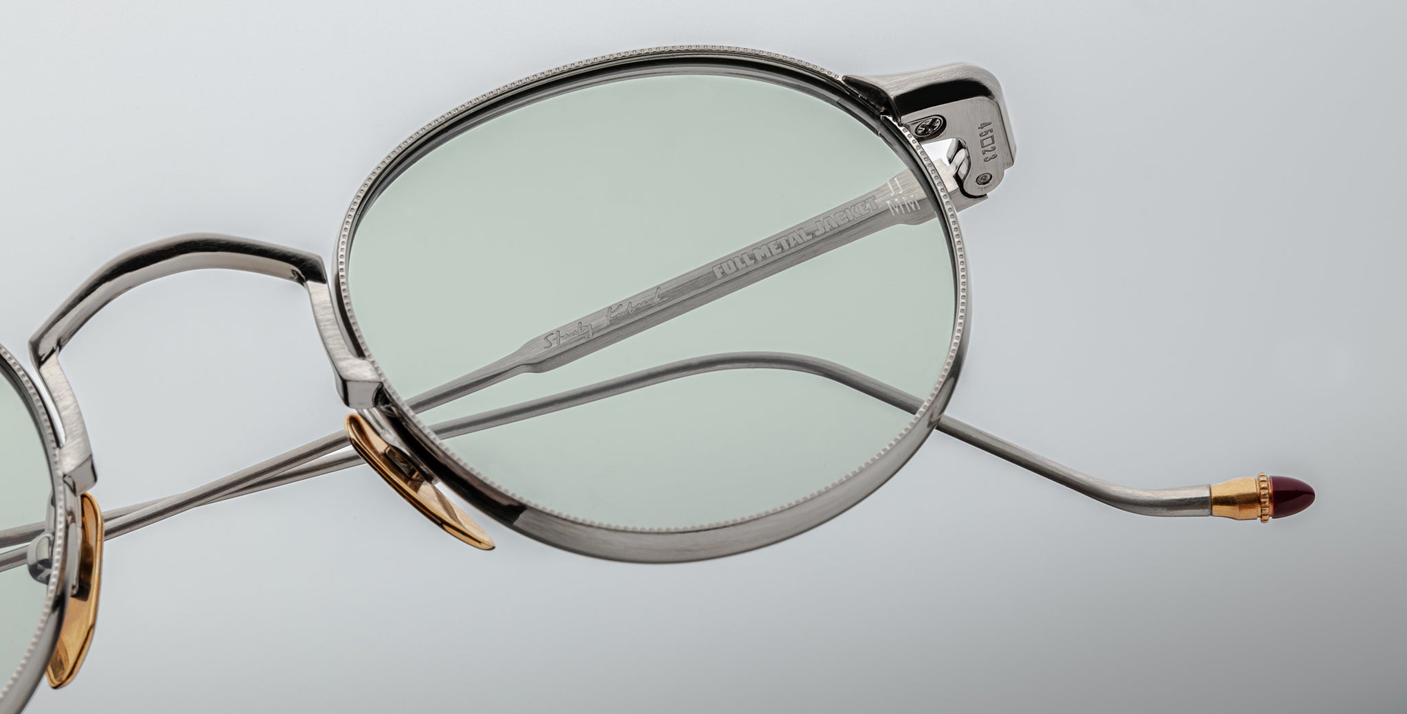Close-up of Jacques Marie Mage Full Metal Jacket round silver metal eyeglasses featuring thin frames, gold nose pads, and a red-tipped temple arm against a light gray background.