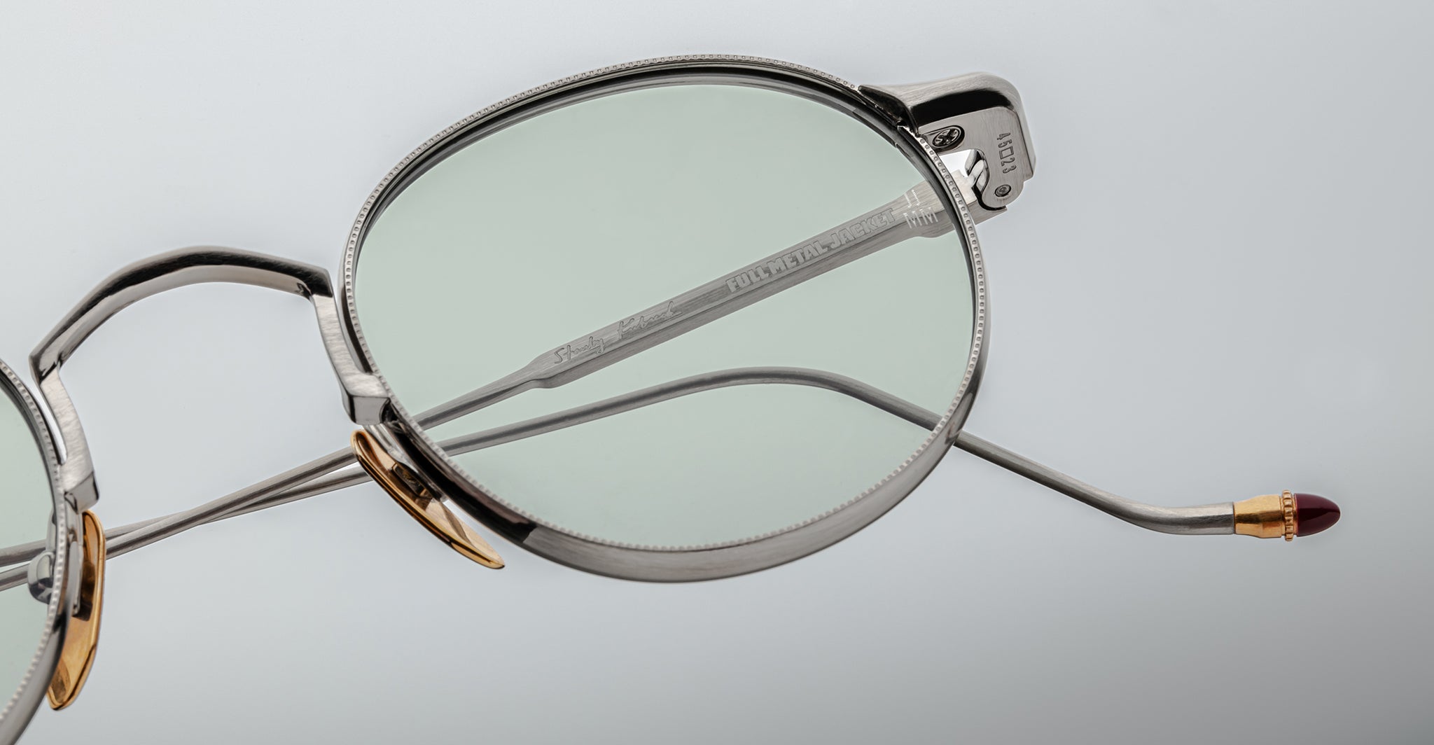 Close-up of Jacques Marie Mage Full Metal Jacket round silver metal eyeglasses featuring thin frames, gold nose pads, and a red-tipped temple arm against a light gray background.