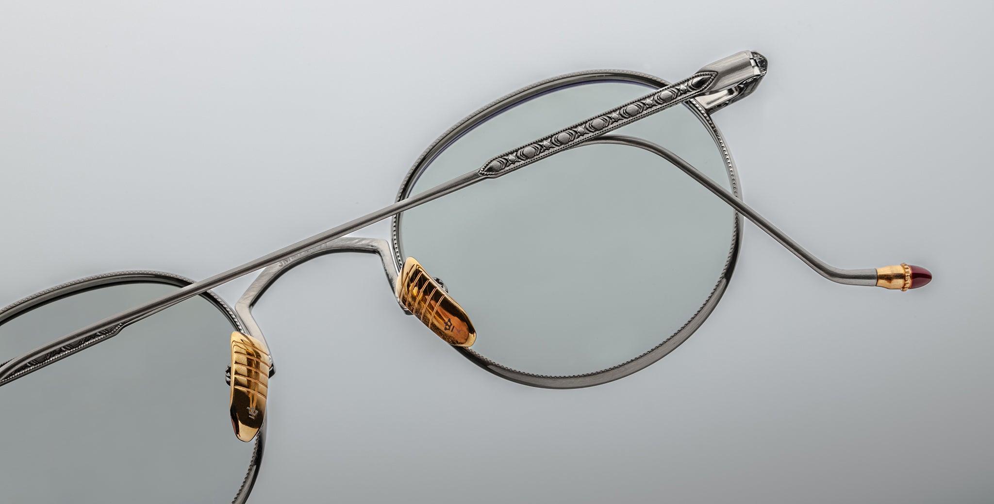 Close-up of Jacques Marie Mage Full Metal Jacket eyeglasses featuring silver metal round lenses, gold nose pads, a single extended temple arm with fine detailing, and a burgundy tip against a light gray background.