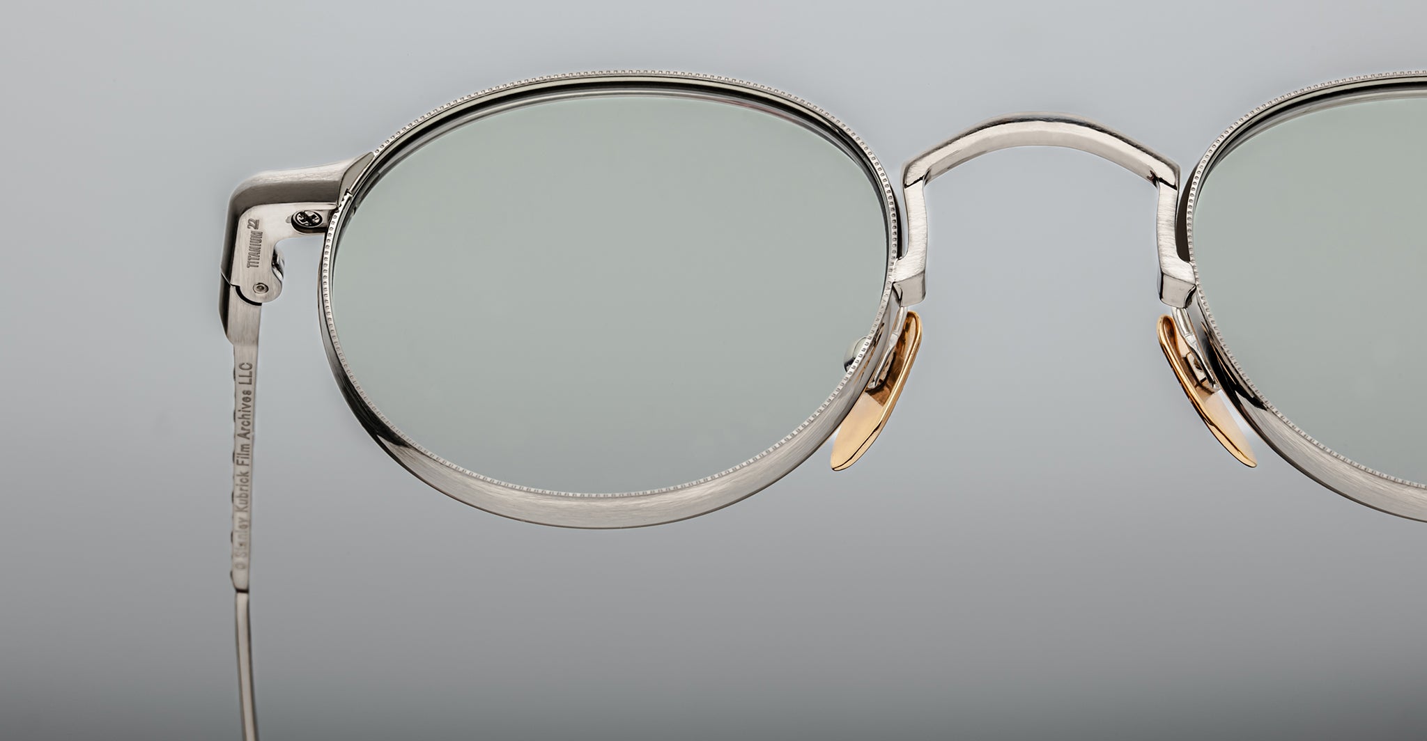 Close-up of Jacques Marie Mage Full Metal Jacket eyeglasses featuring silver oval thin metal frames, clear lenses, and gold nose pads set against a plain, light gray background.
