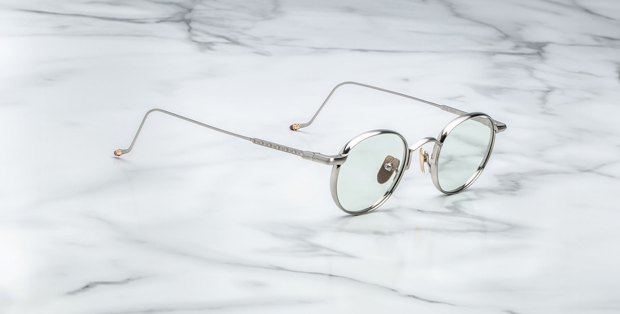 The Jacques Marie Mage Full Metal Jacket features round, silver metal frames with thin arms and clear lenses, displayed on a white marble surface.