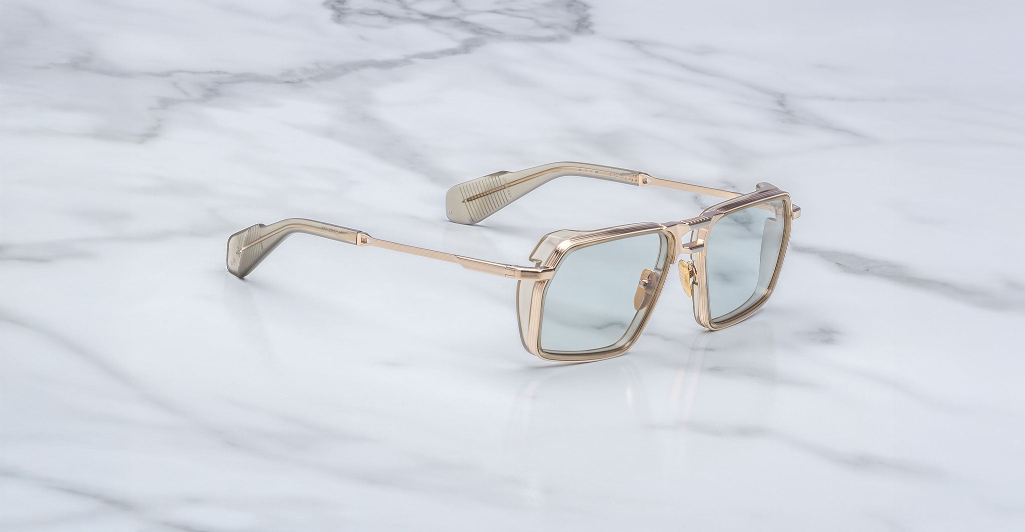 The Jacques Marie Mage GT-R sunglasses feature gold frames, light blue tinted lenses, thick temples, and a modern geometric design, shown resting on a white marble surface.