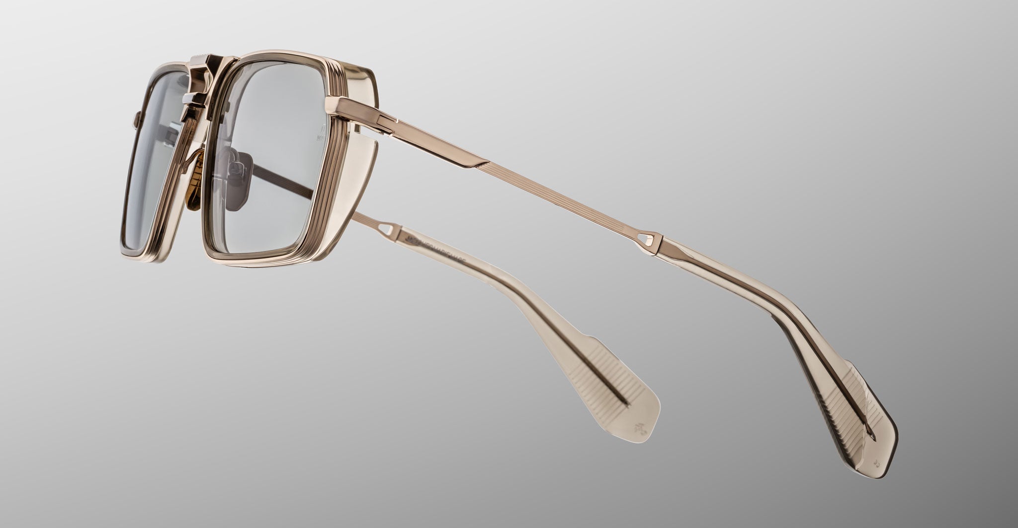 The Jacques Marie Mage GT-R eyeglasses feature a gold metal rectangular frame with clear lenses and detailed temple arms, displayed from a side angle against a light gray background.