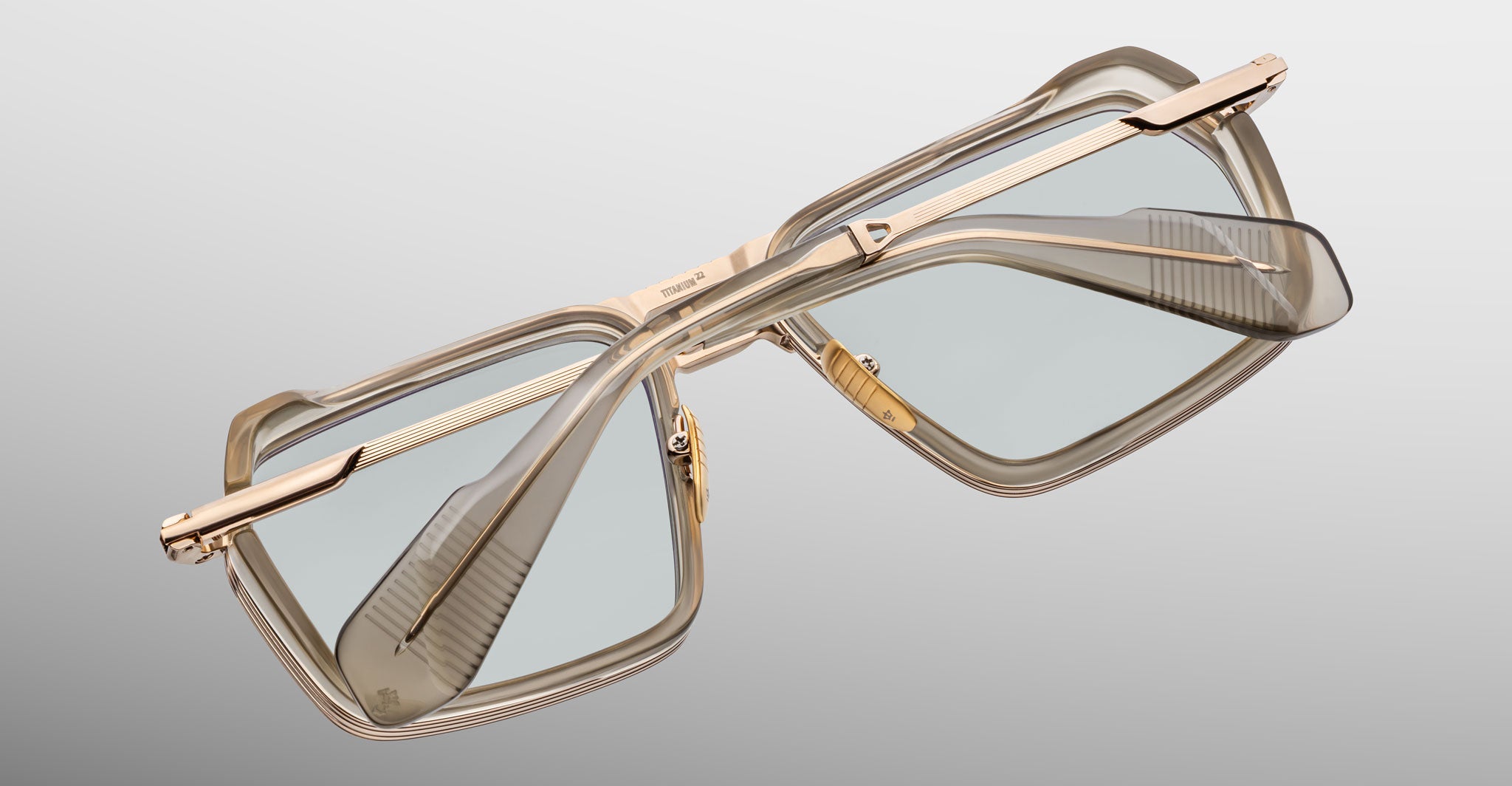 The Jacques Marie Mage GT-R features rectangular eyeglasses with translucent beige frames, gold metal temples, and lightly tinted lenses, displayed at an angle on a neutral grey background.