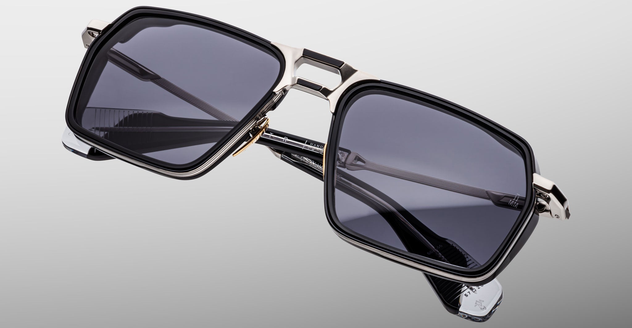 The Jacques Marie Mage GT-R features rectangular black frames, dark tinted lenses, and metallic accents on the bridge and temples, showcased against a neutral gradient background.
