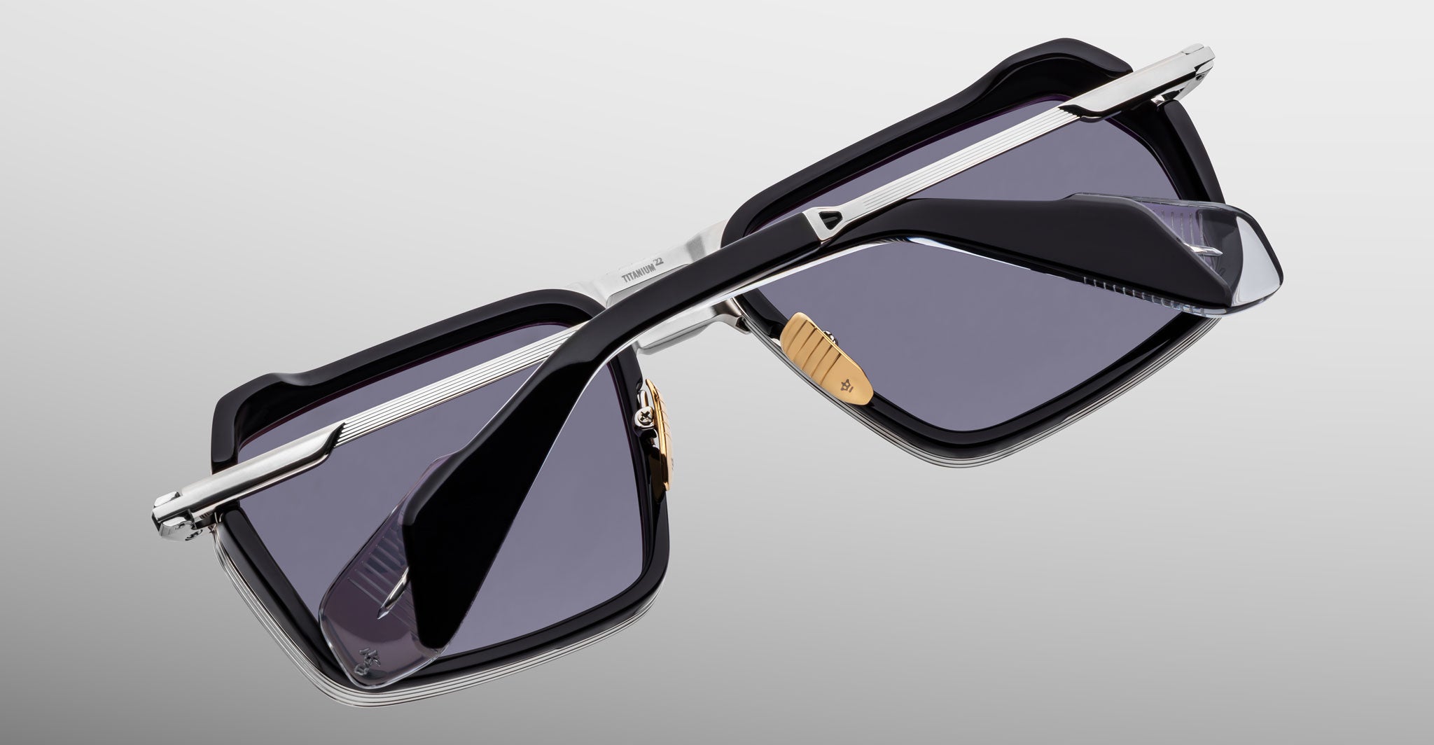 The Jacques Marie Mage GT-R sunglasses feature a black rectangular frame, silver metal arms, and dark tinted lenses, shown resting upside down on a neutral gradient background.