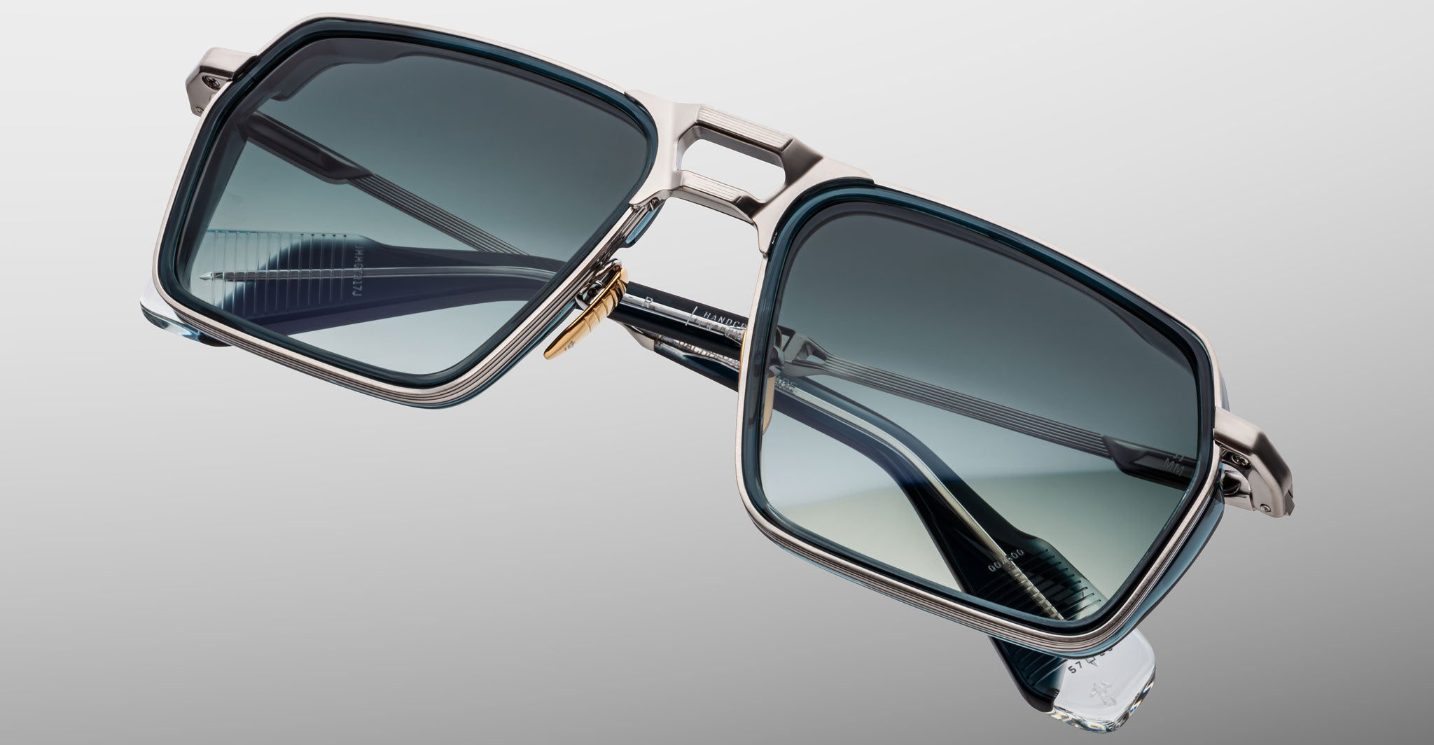 A close-up of Jacques Marie Mage GT-R sunglasses, featuring modern square metallic frames and dark gradient lenses, set against a plain gray background.