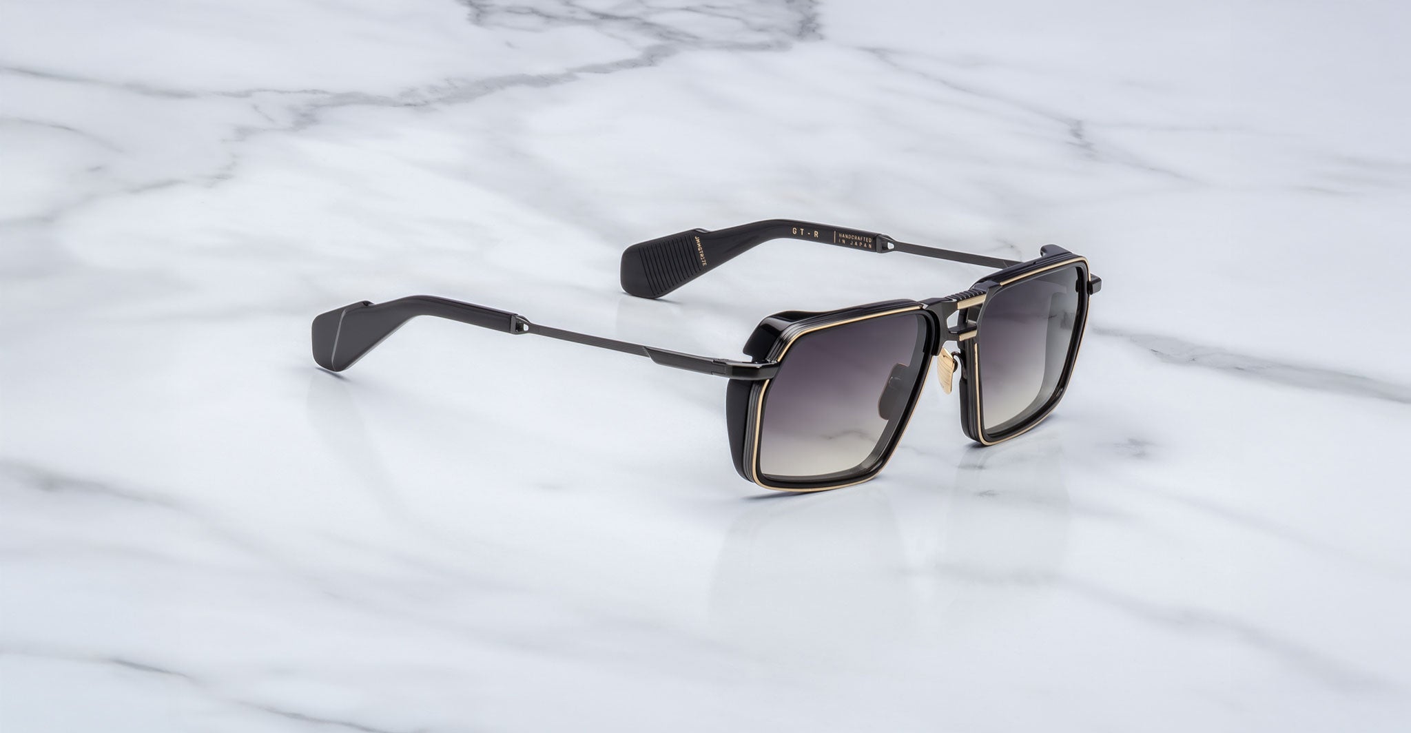 A pair of Jacques Marie Mage GT-R black square sunglasses with gold trim and dark gradient lenses rests on a white marble surface.