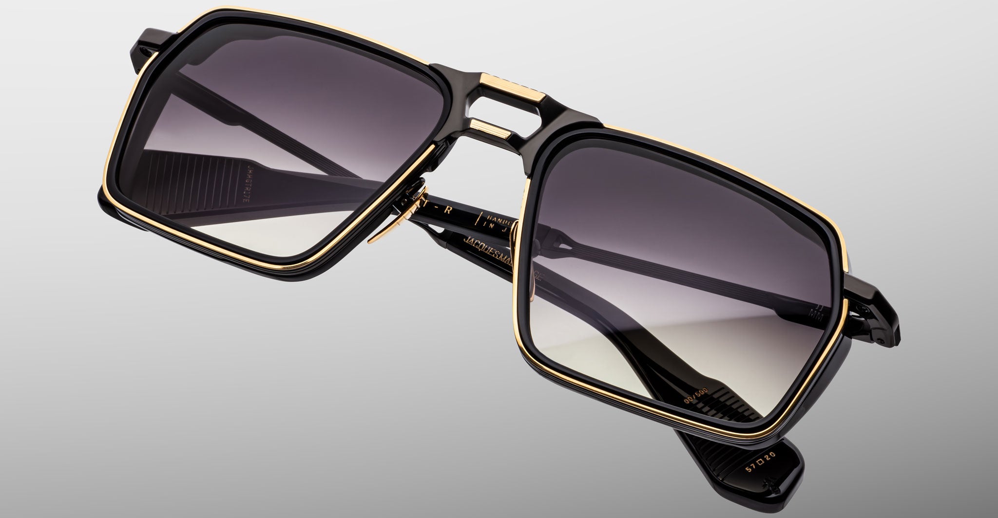 A close-up of Jacques Marie Mage GT-R sunglasses showcases black rectangular frames with gold accents and gradient dark lenses, angled against a plain light gray background.